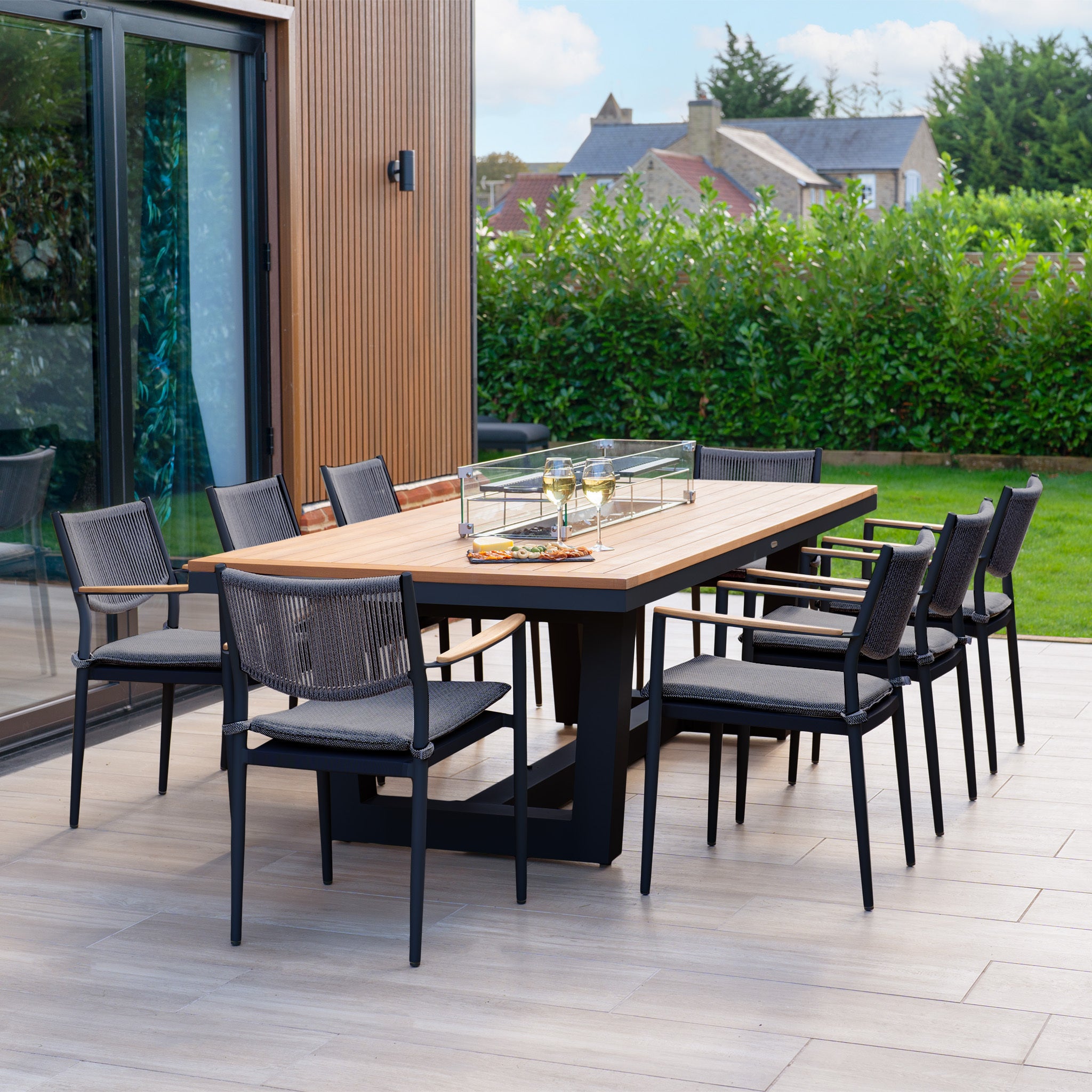 The patio showcases the Tellaro 8 Seat Rectangular Teak Firepit Dining Set in Charcoal, complete with a teak table and eight chairs featuring weather-resistant rope backrests, beautifully surrounded by lush greenery.