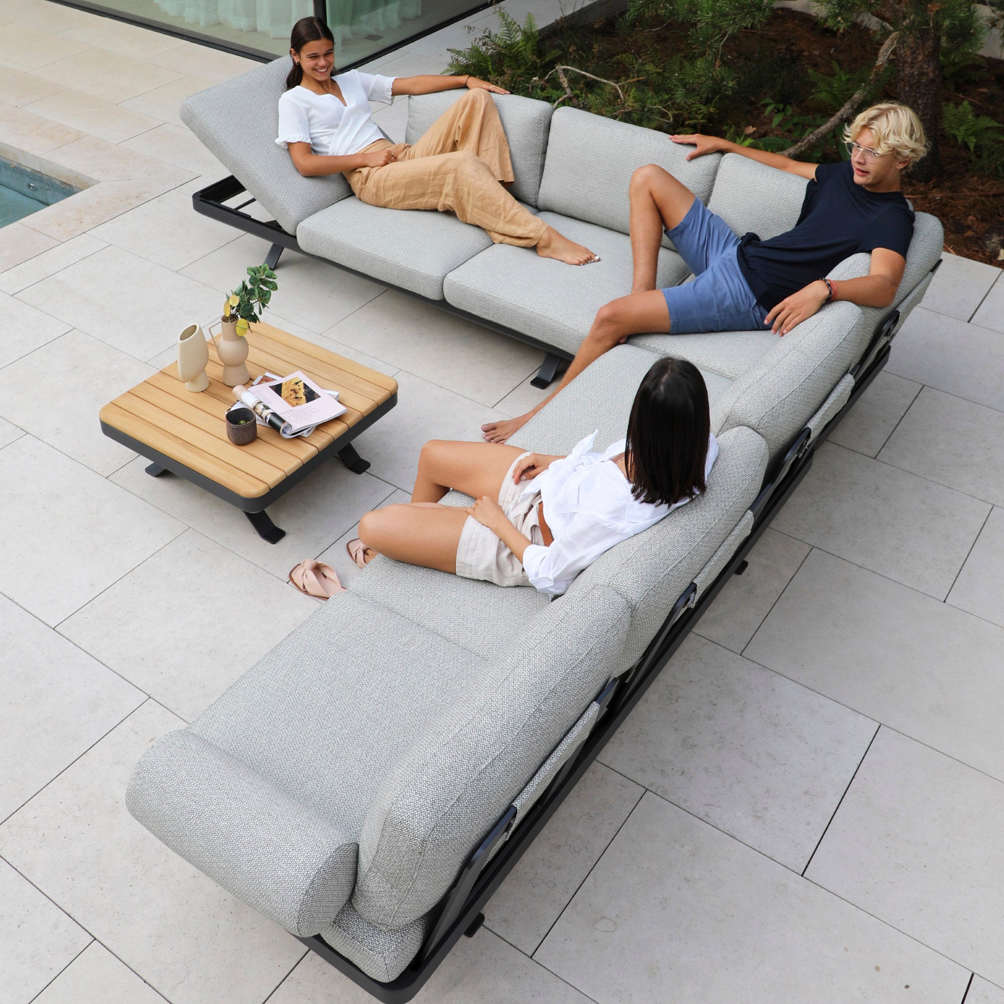 Three people relax on a spacious Vienna Corner Group Set in Grey near a pool, with a wooden coffee table holding a teapot and cups, epitomizing high-end materials and outdoor living.