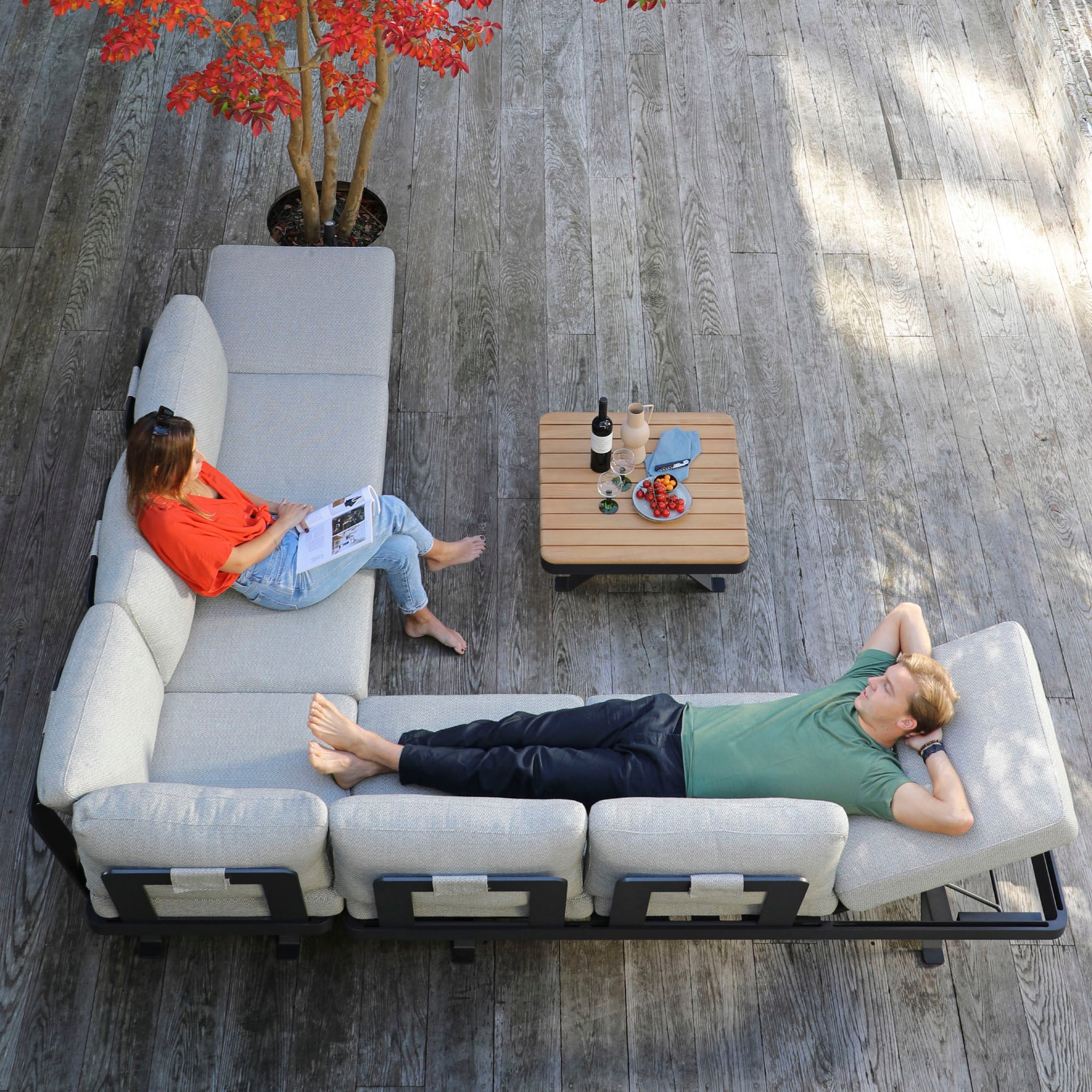 Two people relaxing on an outdoor sectional sofa, enjoying their Vienna Corner Group Set in Grey. The table between them is set with wine, snacks, and a book—perfect for a stylish afternoon of outdoor living with high-end materials.