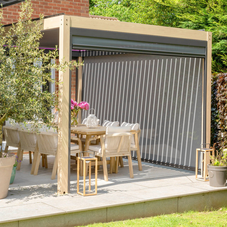 Outdoor dining area with a wooden table, white chairs, a PergoSTET 3m x 3m Square Pergola with 3 Drop Sides and LED Lighting featuring retractable curtains and a waterproof roof, surrounded by lush greenery.