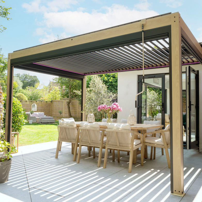 Outdoor dining area under a contemporary PergoSTET 3m x 4m Rectangular Pergola with 3 Drop Sides and LED Lighting with Wood Effect, featuring a wooden table, chairs, and flowers. Adjacent to the garden with patio furniture, this setup offers an inviting ambiance enhanced by the stylish and shaded pergola.