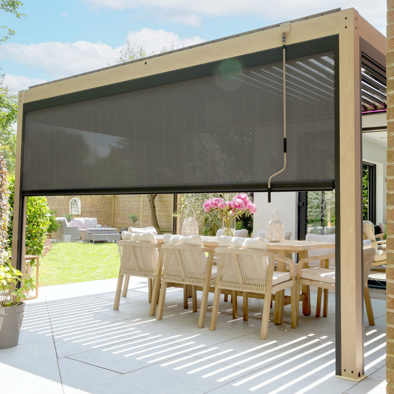 Covered outdoor dining area with a wooden table and chairs, shaded by a PergoSTET 3m x 4m Rectangular Pergola with 3 Drop Sides and LED Lighting featuring a wood effect, surrounded by greenery on a sunny day.