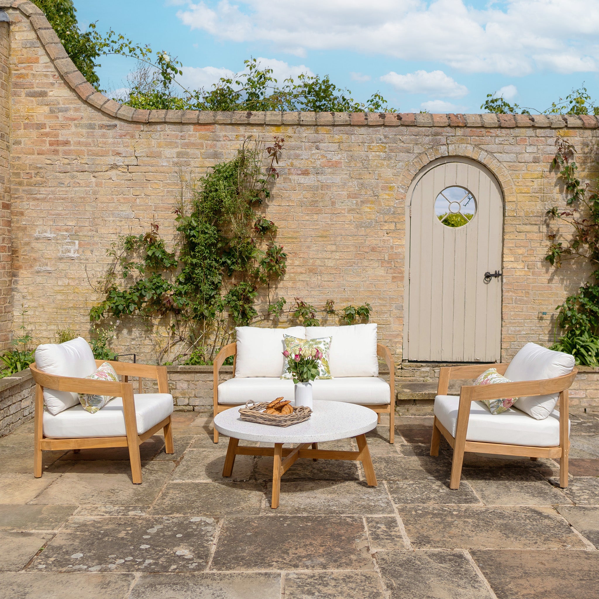 The Yosemite 2 Seat Sofa Set In Glacier includes white cushions, wooden chairs, a loveseat, and a round table made of premium teak wood. Set against a backdrop of brick wall with greenery, the cushions are crafted from water-resistant polyester fabric for enhanced durability.