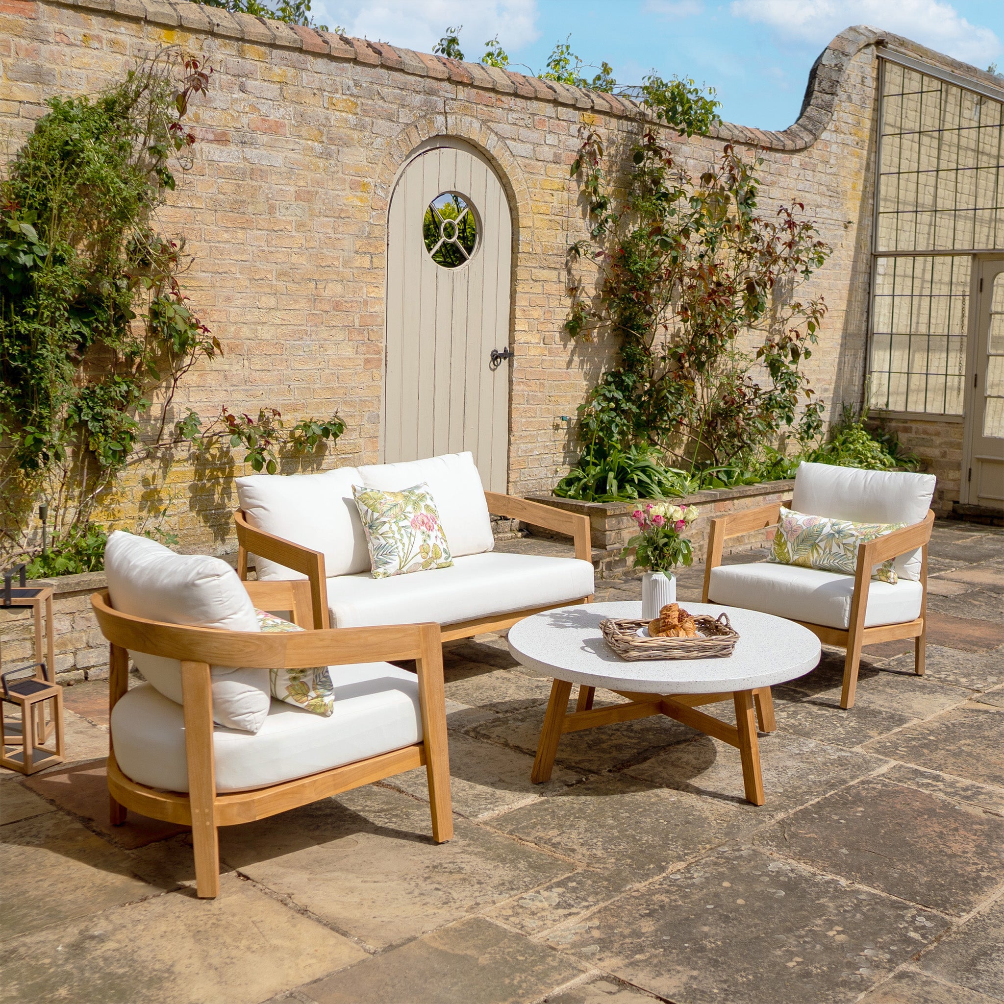 Outdoor patio featuring the Yosemite 2 Seat Sofa Set in Glacier, adorned with premium teak wood and white water-resistant polyester cushions, accompanied by a round table with pastries, all set against a brick wall backdrop.