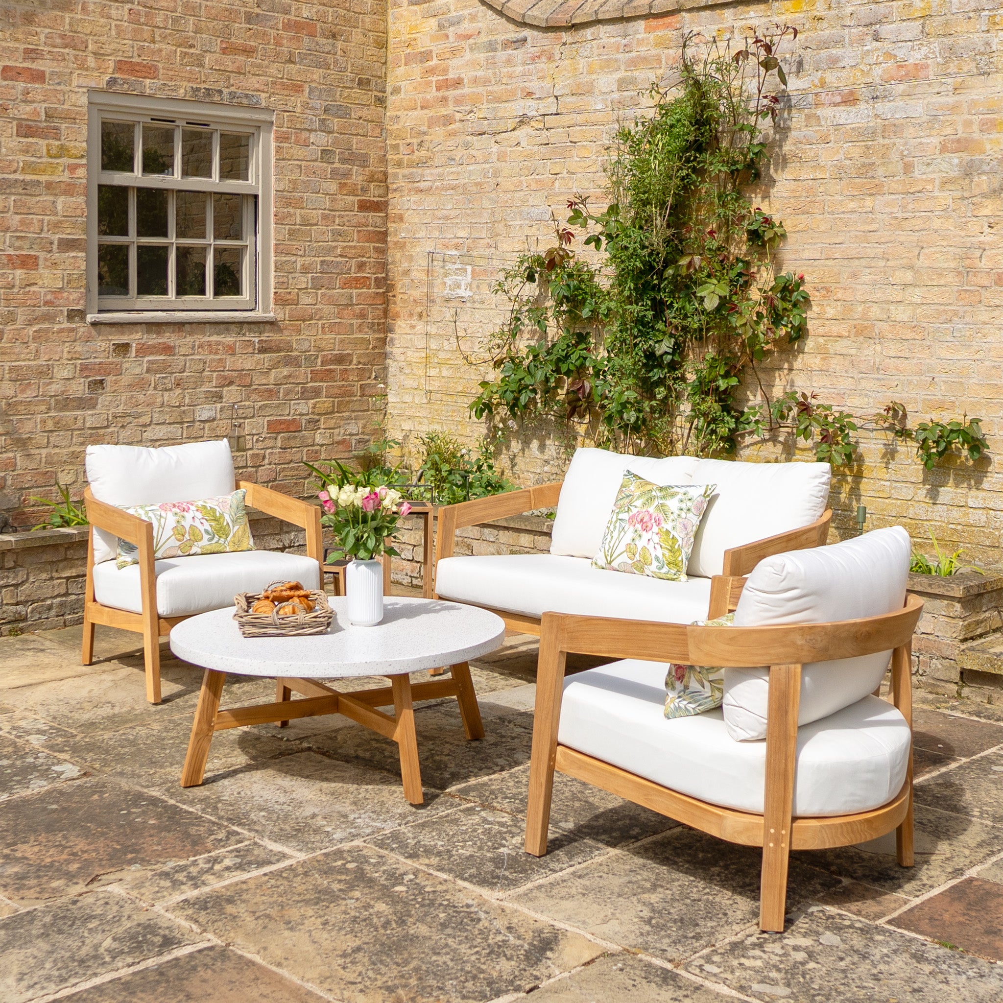 Outdoor patio with the Yosemite 2 Seat Sofa Set In Glacier, featuring premium teak wood furniture, white cushions made from water-resistant polyester fabric, a coffee table adorned with flowers and snacks, and a vine-covered brick wall.