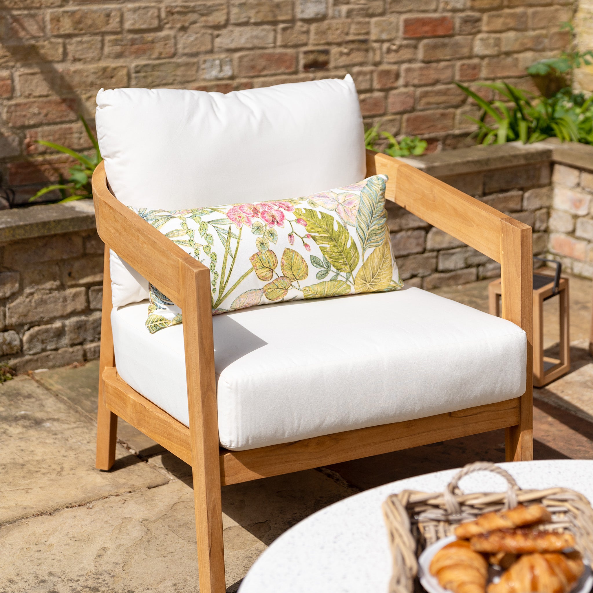 A Yosemite 2 Seat Sofa Set In Glacier, featuring white cushions and a floral pillow made of water-resistant polyester fabric, sits next to a table with pastries on a sunny patio.