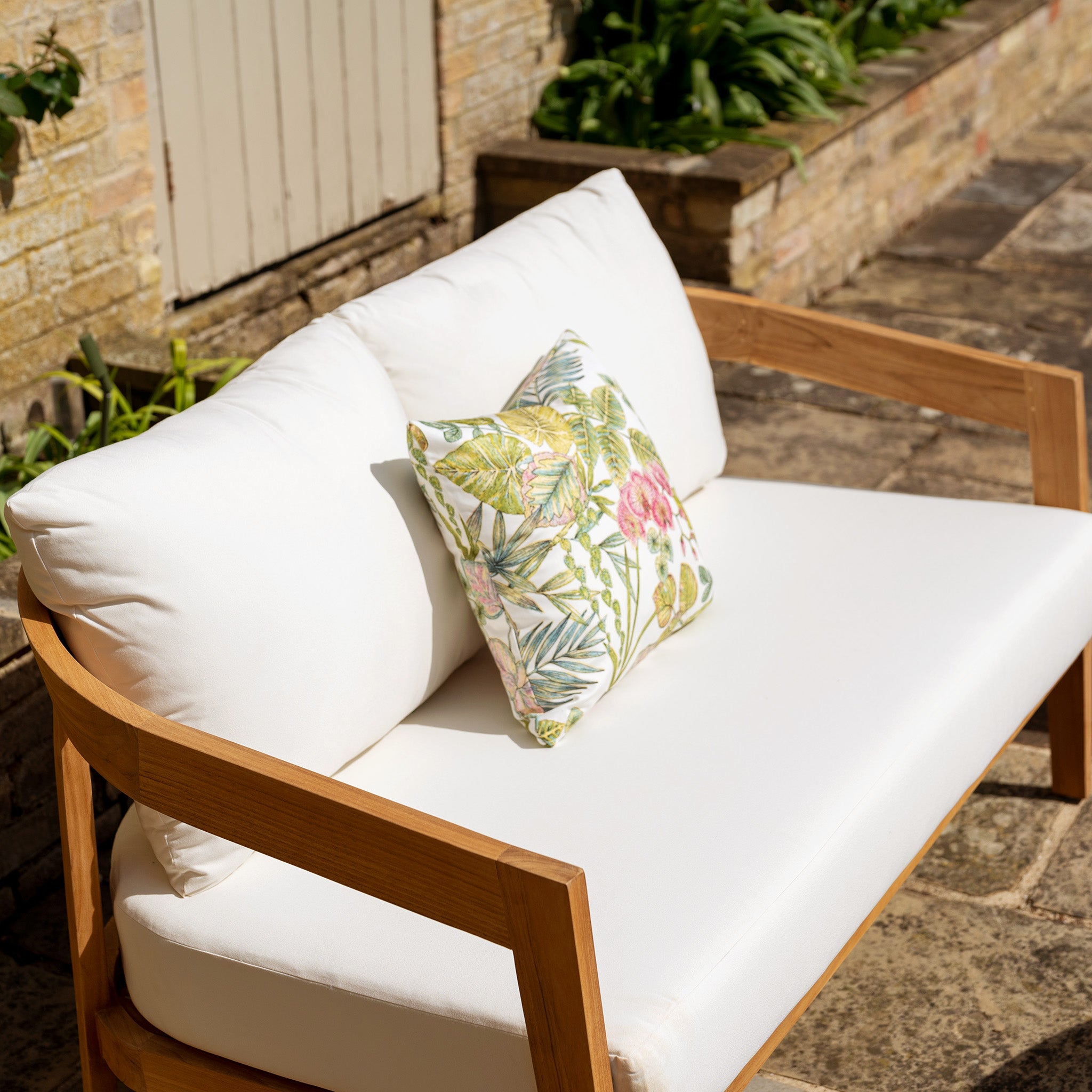 A Yosemite 2 Seat Sofa Set in Glacier, adorned with a floral pillow, rests on a stone patio near a brick wall with plants. The cushions, crafted from water-resistant polyester fabric, ensure both durability and comfort.