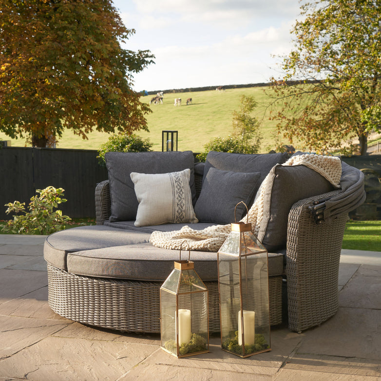 The Pacific Lifestyle Bermuda Daybed in Slate Grey offers a space-saving design with a round wicker lounge, cushions, and two weatherproof lanterns, perfect for enjoying countryside views with grazing cows.