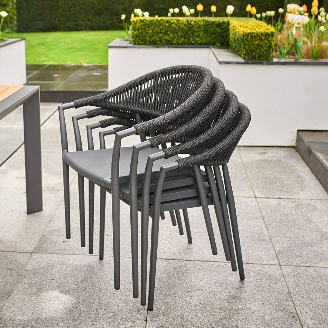 Stacked black woven chairs on a grey patio with a garden and flowers in the background, perfectly complementing the Cloverly 8 Seat Rectangular Extending Dining Set with Teak Table in Charcoal.