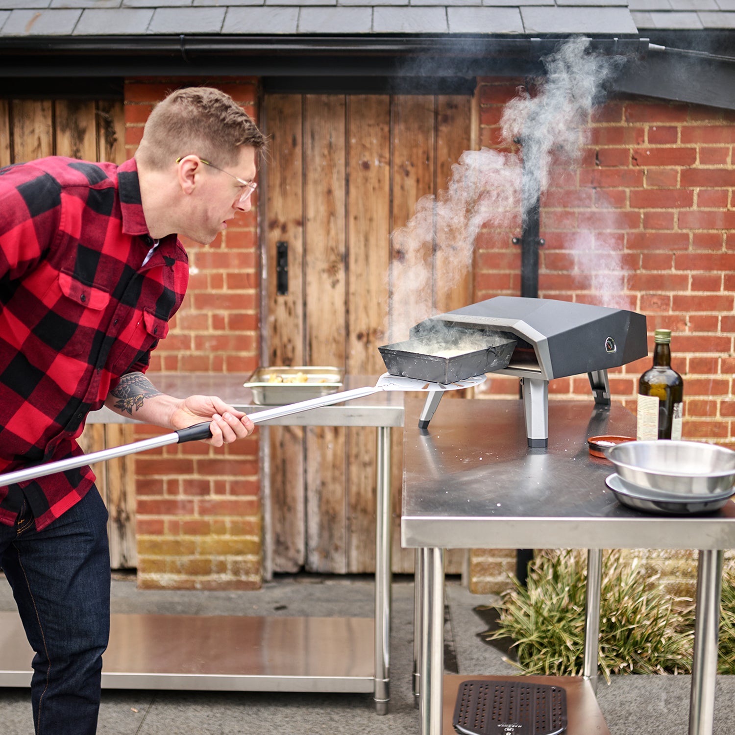 Person using a metal peel to insert or remove stone-baked pizza from a smoking Juno 12" Pellet Pizza Oven, with a brick wall in the background.