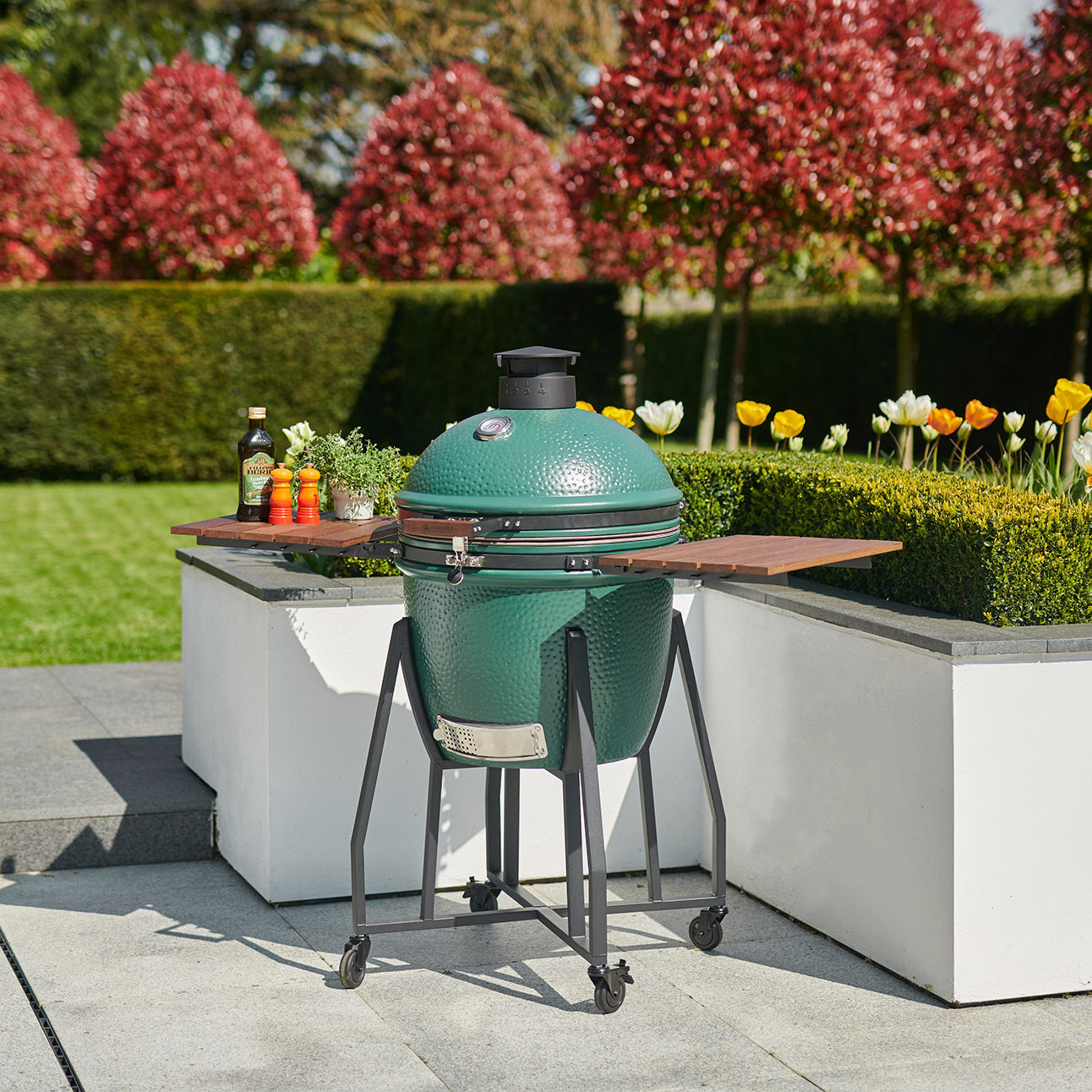 A Kamado BBQ 18" Ceramic Grill in Matte Green, featuring ceramic walls and mounted on a stand with wooden shelves, nestled in a garden with manicured hedges, tulips, and red-leaved trees.