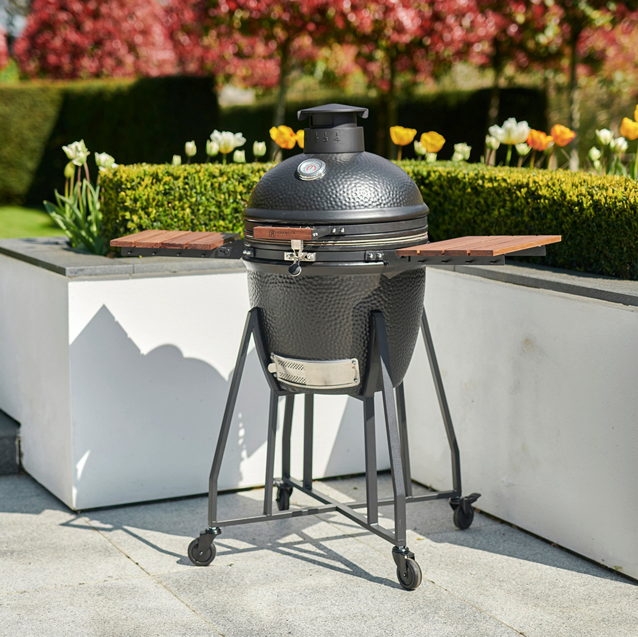 A Kamado BBQ 18" Ceramic Grill in Black, featuring a round lid and wooden side shelves on a wheeled stand, is the perfect addition to any garden setup.