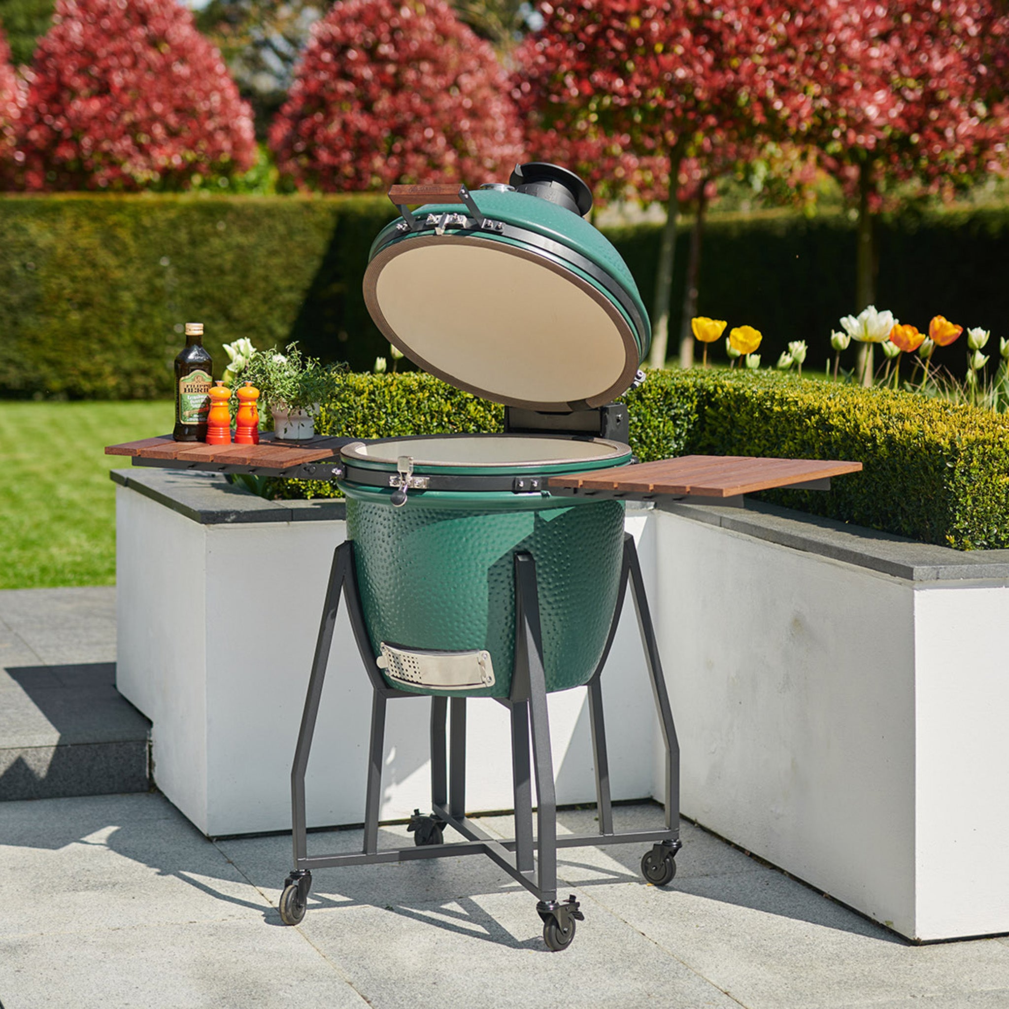 Kamado BBQ 18" Ceramic Grill in Matte Green on a wheeled stand with an open lid, set in a garden with tulips, shrubs, and bottles on adjacent wood shelves.