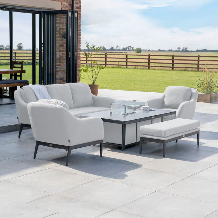 Modern outdoor patio set composed of the Luna 3 Seat Outdoor Fabric Sofa Set with Rising Firepit Table in Oyster Grey, featuring plush white cushions on a spacious tiled patio overlooking a field.