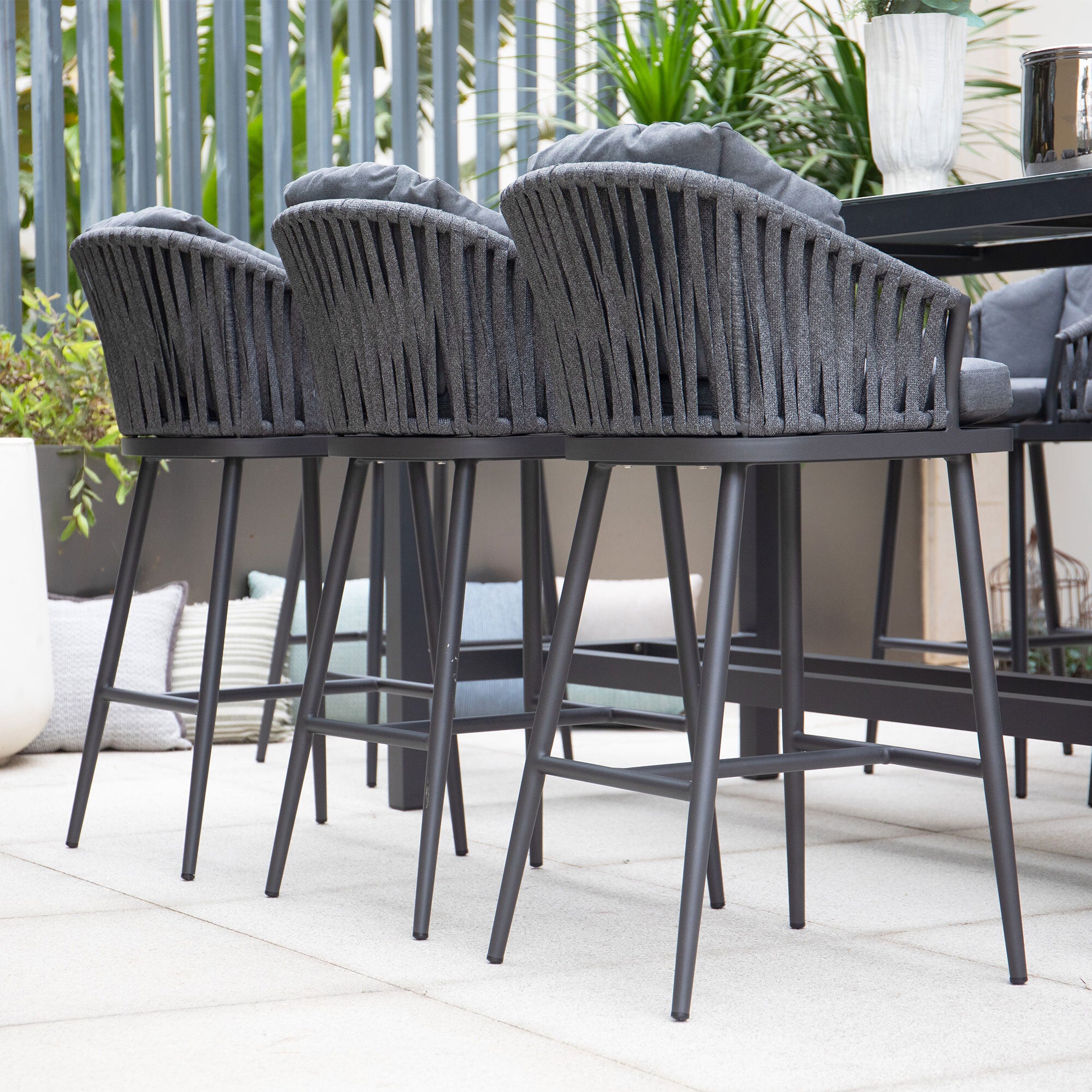 Three Monterrey Bar Stools in Grey arranged at a high table on a patio adorned with potted plants, creating an ideal setting for your UV-resistant outdoor living area.