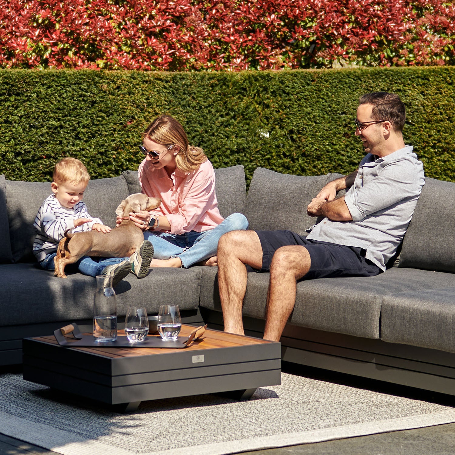 A family of three, along with their small dog, sits on an outdoor sofa in a garden, smiling and playing together. Their Panama Luxury Outdoor L-Shape Corner Group Set in Charcoal, crafted from sustainable teak by Harbour Lifestyle, adds a touch of luxury to their outdoor oasis.