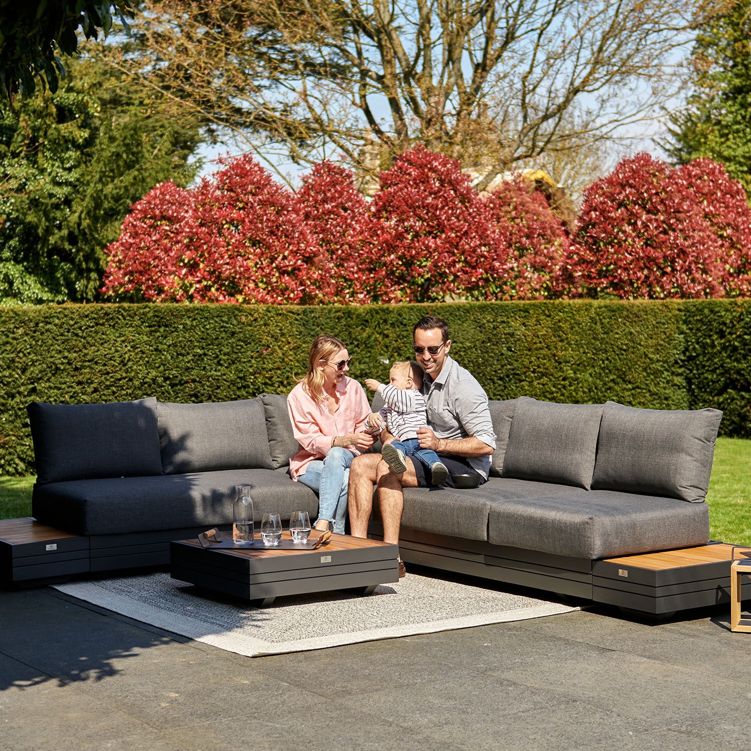 A family sits on their Panama Luxury Outdoor L-Shape Corner Group Set in Charcoal with a child, surrounded by greenery and a vibrant hedge in the background, enjoying their Harbour Lifestyle piece made from sustainable teak.