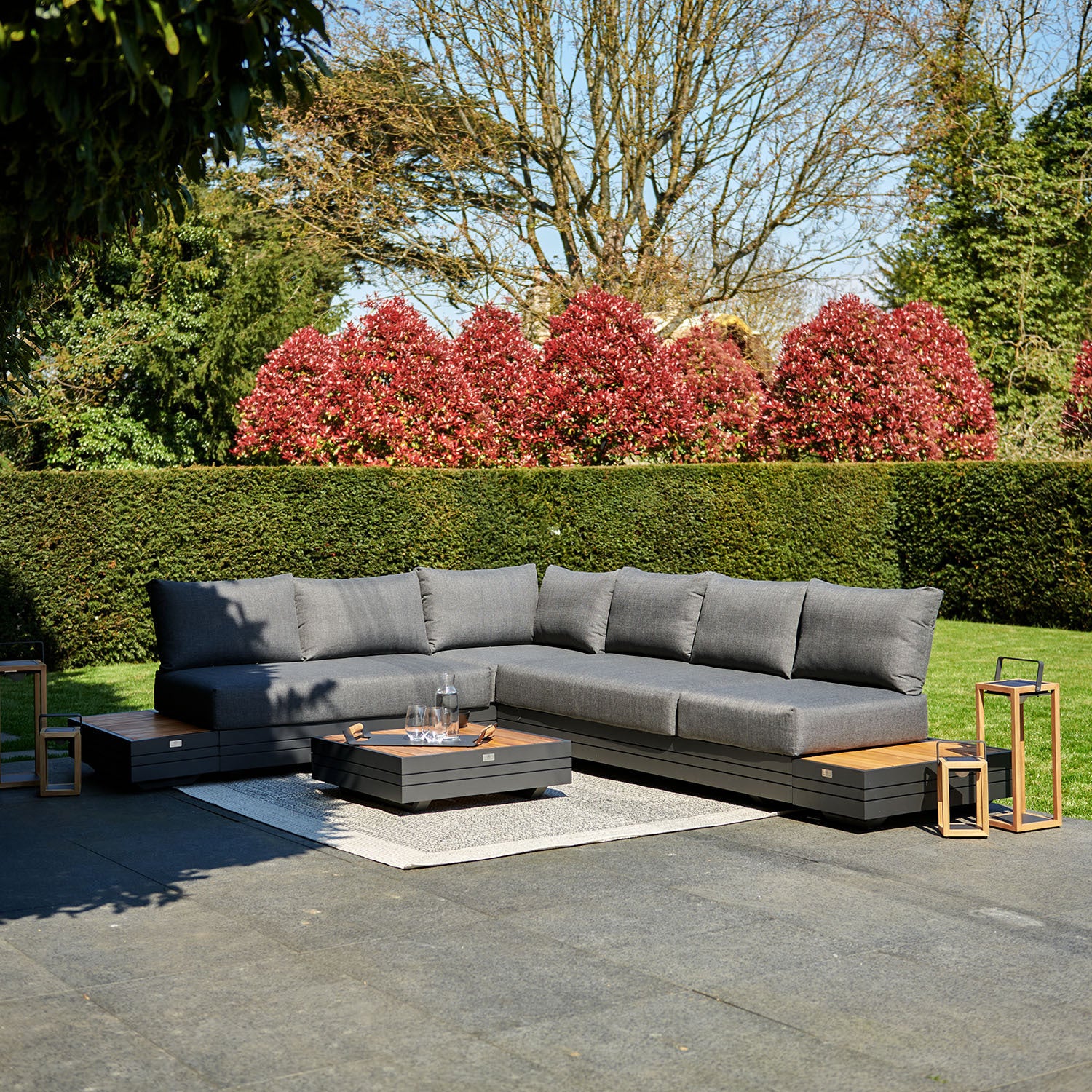 A modern outdoor seating area with a charcoal sectional sofa, low table, lanterns, and a manicured garden in the background showcases the Panama Luxury Outdoor L-Shape Corner Group Set in Charcoal from Harbour Lifestyle, crafted from sustainable teak.