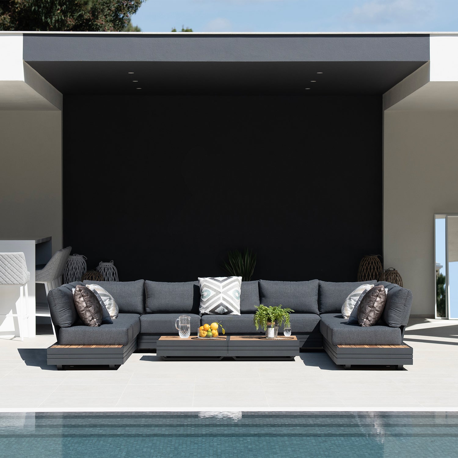 Modern outdoor patio featuring a Panama Luxury Outdoor Large U-Shape Corner Sofa Set in Charcoal by Harbour Lifestyle, adorned with patterned pillows. A centerpiece showcases drinks, fruit, and a plant for relaxed vibes. Sustainable teak accents complete the luxurious look.