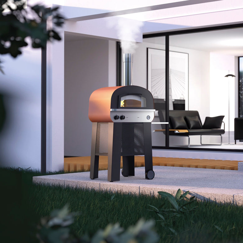 Fontana Piero Gas & Wood Pizza Oven, complete with trolley and chimney, positioned on a patio in front of a contemporary house with glass sliding doors.