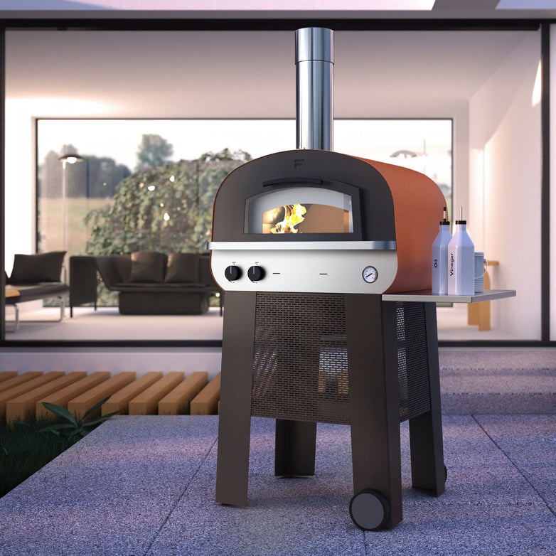 The Fontana Piero Gas & Wood Pizza Oven Including Trolley warms the modern patio with a fire burning inside, while a cozy seating area and large window provide the perfect backdrop.