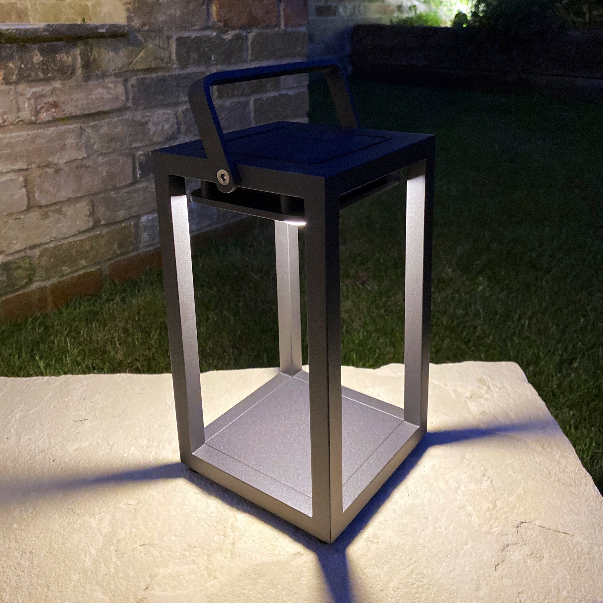 The Luxor Small Table Outdoor and Indoor Solar Lantern in Charcoal, a modern and weatherproof lantern with a handle that emits soft light, is placed on a stone surface outdoors at night.