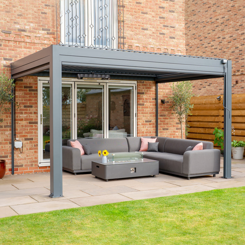 A modern patio with a PergoSTET 3m x 4m Rectangular Wall Mounted Pergola featuring 3 drop sides and LED lighting in gray, a gray sectional sofa, a glass coffee table, and potted plants against a brick house. This sleek setup features a built-in drainage system for added convenience and durability.