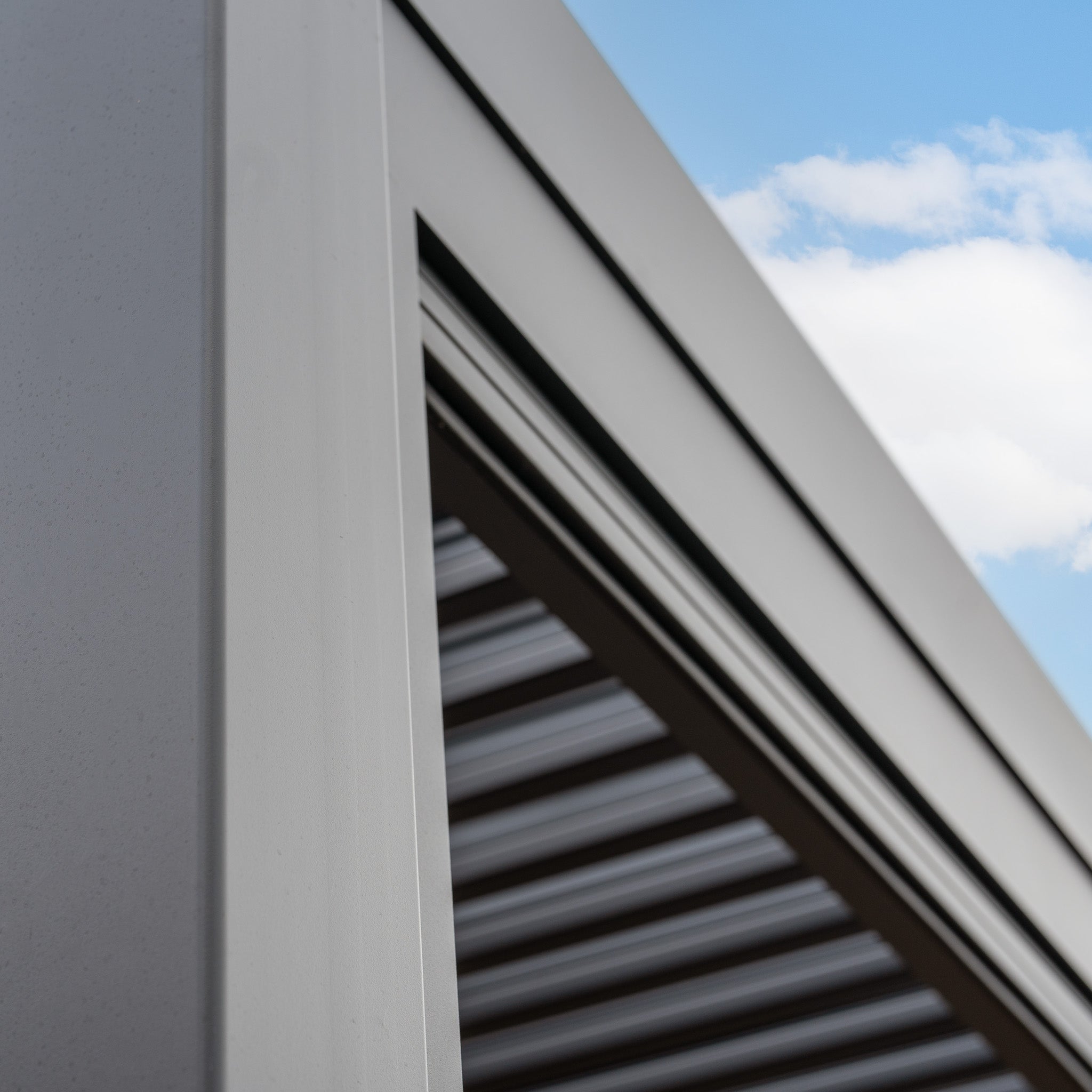 Close-up of a modern PergoSTET 3m x 3m Square Wall Mounted Pergola with 3 Drop Sides and LED Lighting in Grey, featuring a grey metal structure and slatted roof complete with a built-in drainage system, set against a blue sky dotted with scattered clouds.