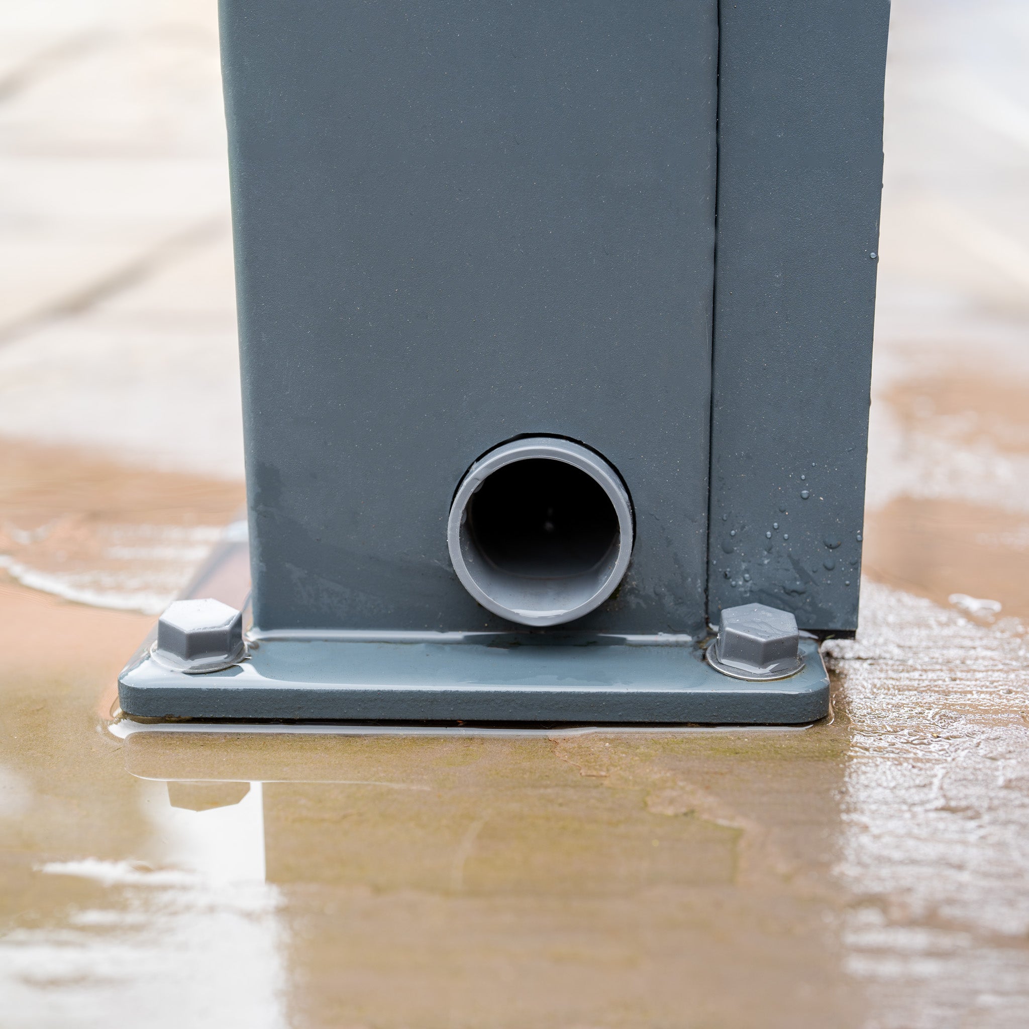 Close-up of a gray metal pole from the PergoSTET 3m x 3m Square Wall Mounted Pergola with 3 Drop Sides and LED Lighting in Grey, featuring a built-in drainage system at its base, water pooling on the surrounding concrete.