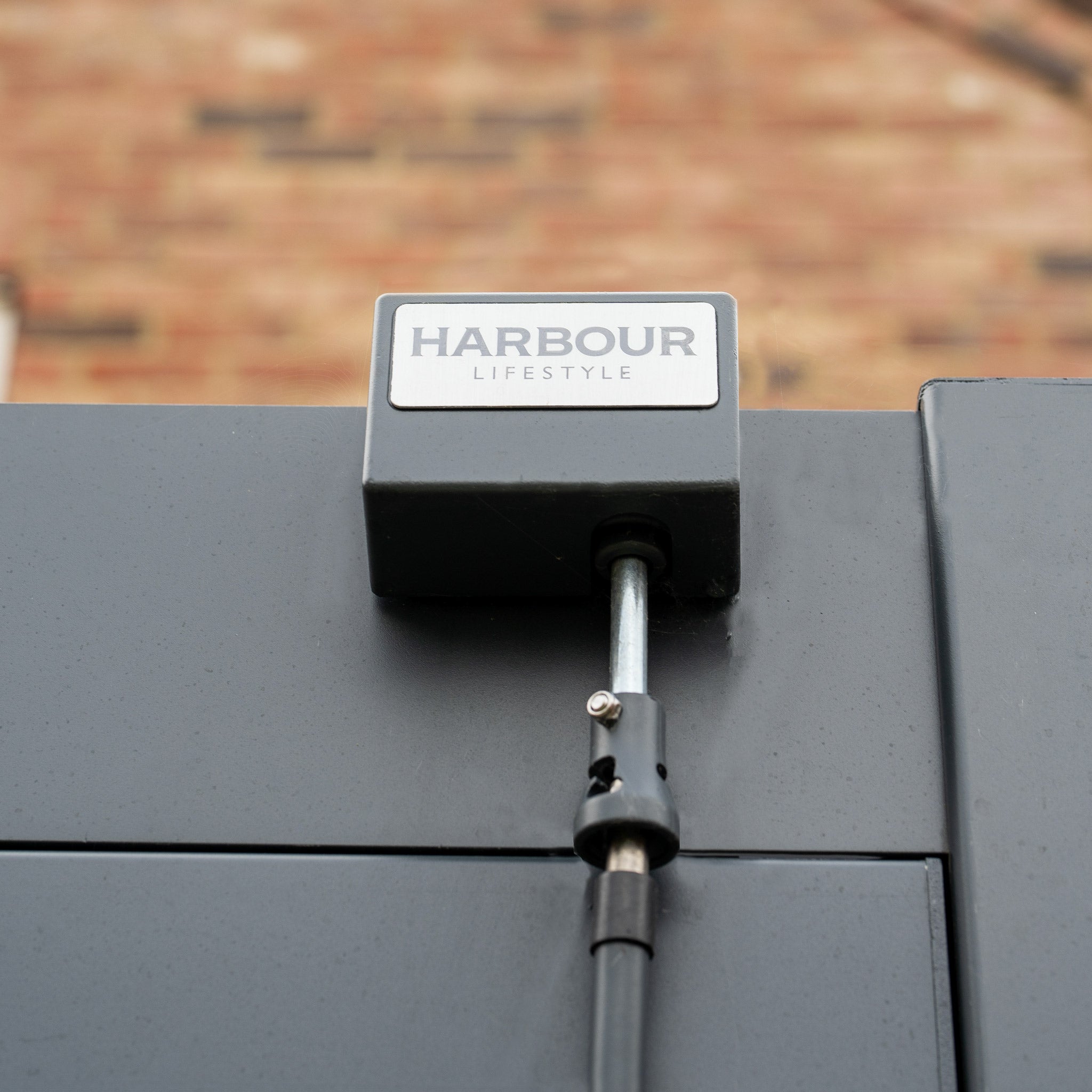 Close-up of a grey fence gate with a metal latch and a sign reading "Harbour Lifestyle" against a brick wall background, perfectly complementing the PergoSTET 3m x 3m Square Wall Mounted Pergola with 3 Drop Sides and LED Lighting in Grey.