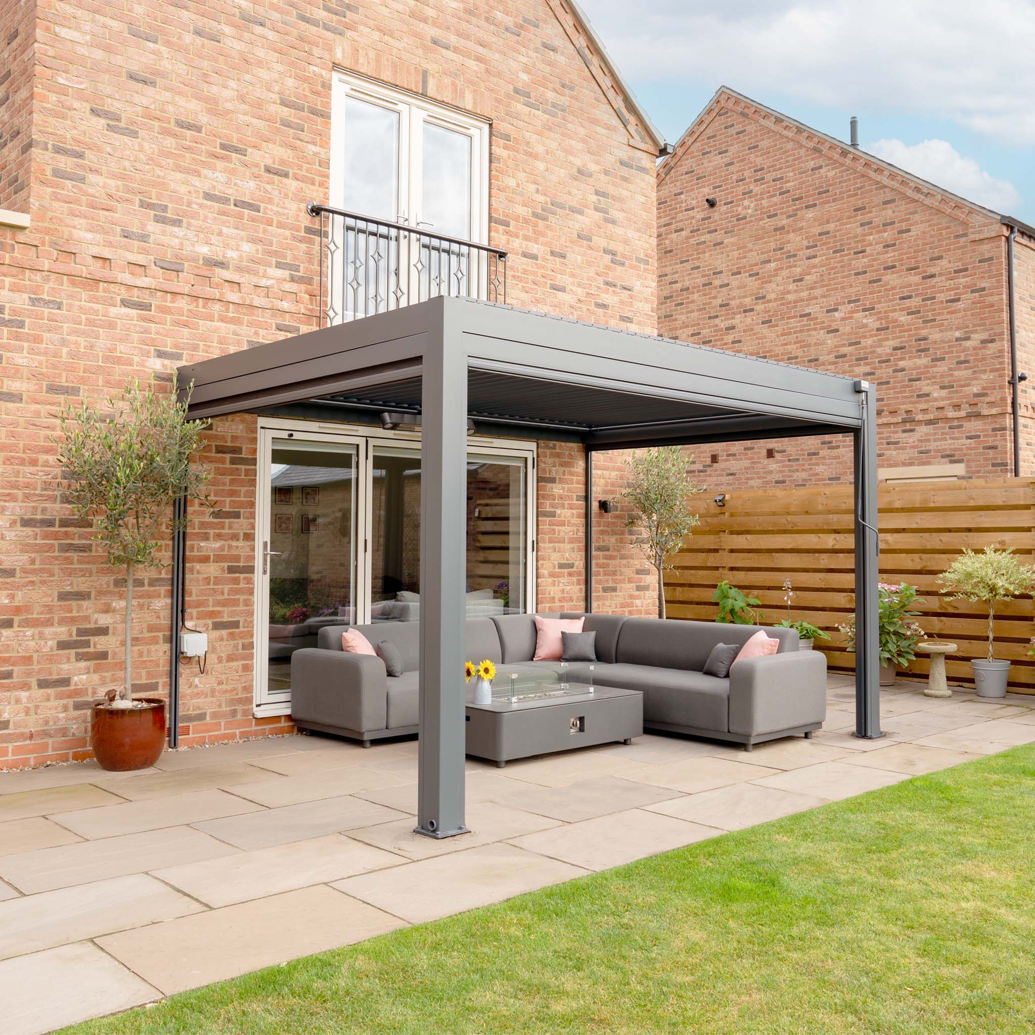 A modern patio featuring a sleek PergoSTET 3m x 4m Rectangular Wall Mounted Pergola with 3 Drop Sides and LED Lighting in Grey, gray outdoor sofas, a glass coffee table, and surrounding plants near the brick house. The area is designed with a built-in drainage system for added functionality.