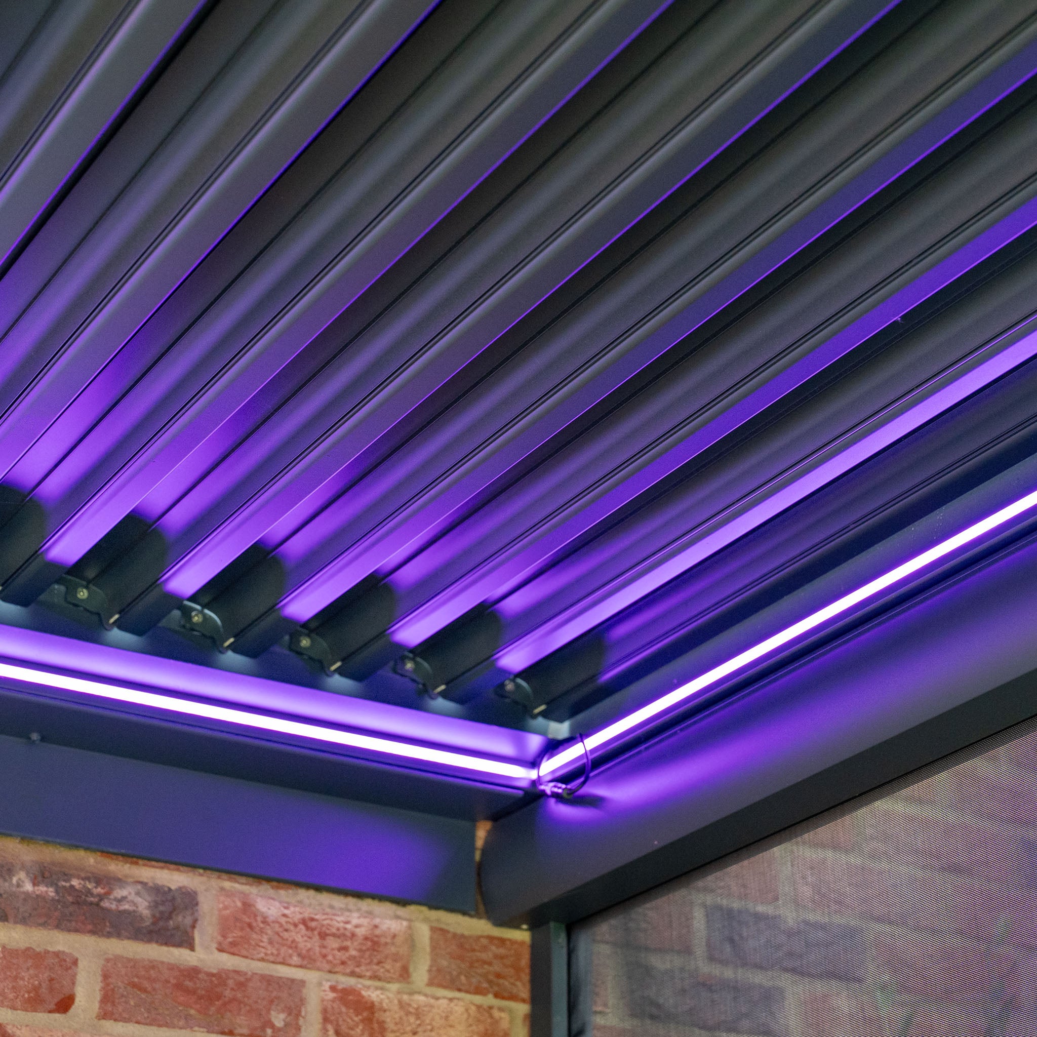 Ceiling featuring angled metal slats and purple LED lighting along the edges, positioned next to a brick wall, reminiscent of the sleek design of the PergoSTET 3m x 3m Square Wall Mounted Pergola with 3 Drop Sides and LED Lighting in Grey.