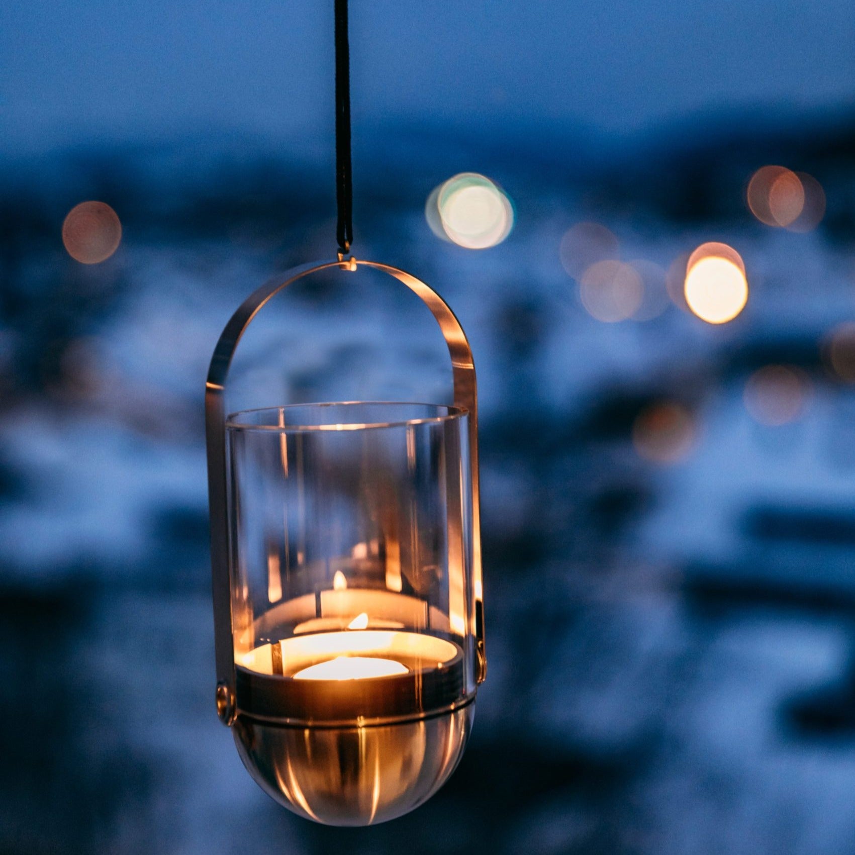 A lit candle inside the Höfats GRAVITY CANDLE Hanger glows warmly against a blurred background of evening lights, suspended elegantly with brushed solid stainless steel elements.