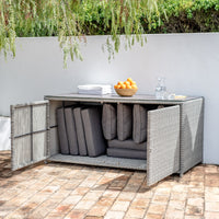 The Santiago Rattan Storage Box/Console Table with Glass Top in Grey on the patio holds folded gray cushions, topped with a bowl of oranges, a water jug, and a glass, serving as both practical storage and an elegant garden accessory.