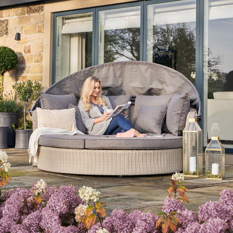 Relaxing on a Pacific Lifestyle Bermuda Daybed in Stone Grey, someone enjoys a book amidst purple flowers and lanterns. The weatherproof material makes this daybed an ideal outdoor retreat.