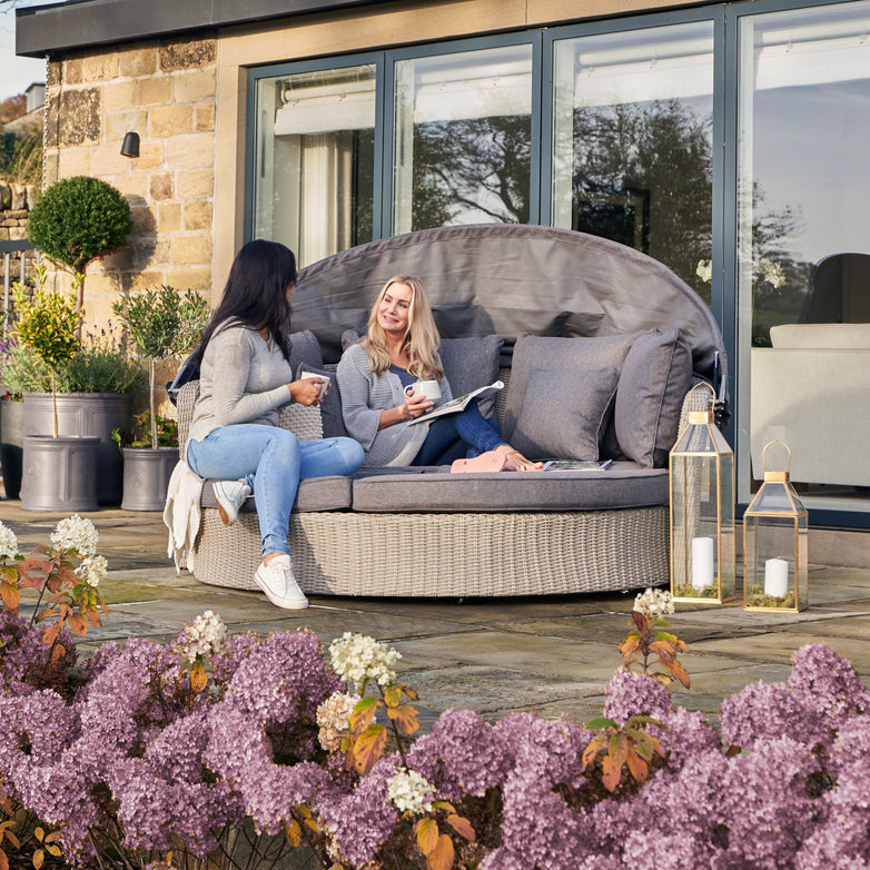 Two women sit on a spacious Pacific Lifestyle Bermuda Daybed in Stone Grey, made from weatherproof material, chatting and holding mugs. Nearby flowers bloom as lanterns cast a warm glow on the patio.