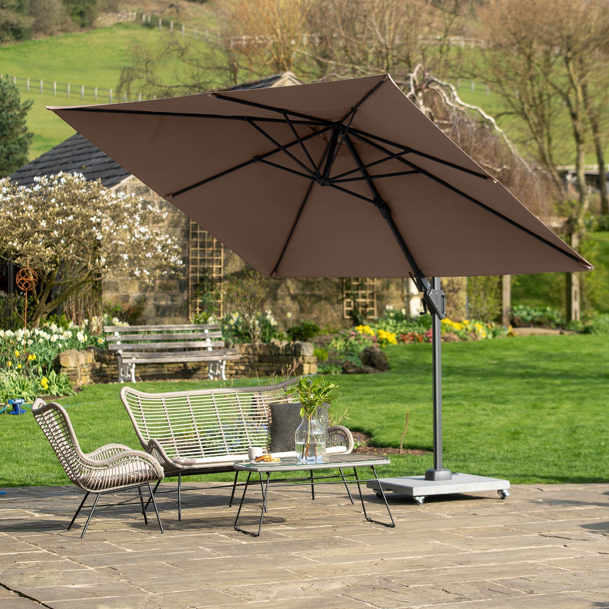 A garden scene with a Voyager T2 2.7m Square Parasol in Taupe shading wicker chairs and a table set on a stone patio, surrounded by lush greenery in the background.