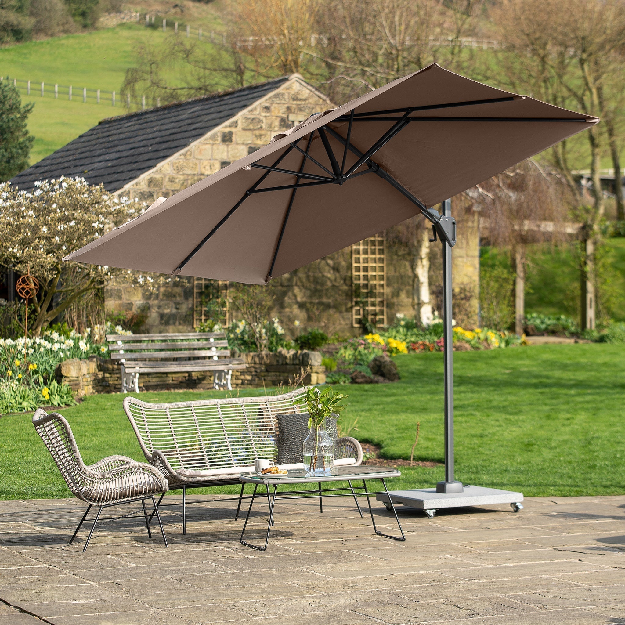 A patio with a large Voyager T2 2.7m Square Parasol in Taupe shading a seating area consisting of a sofa, two chairs, and a coffee table.