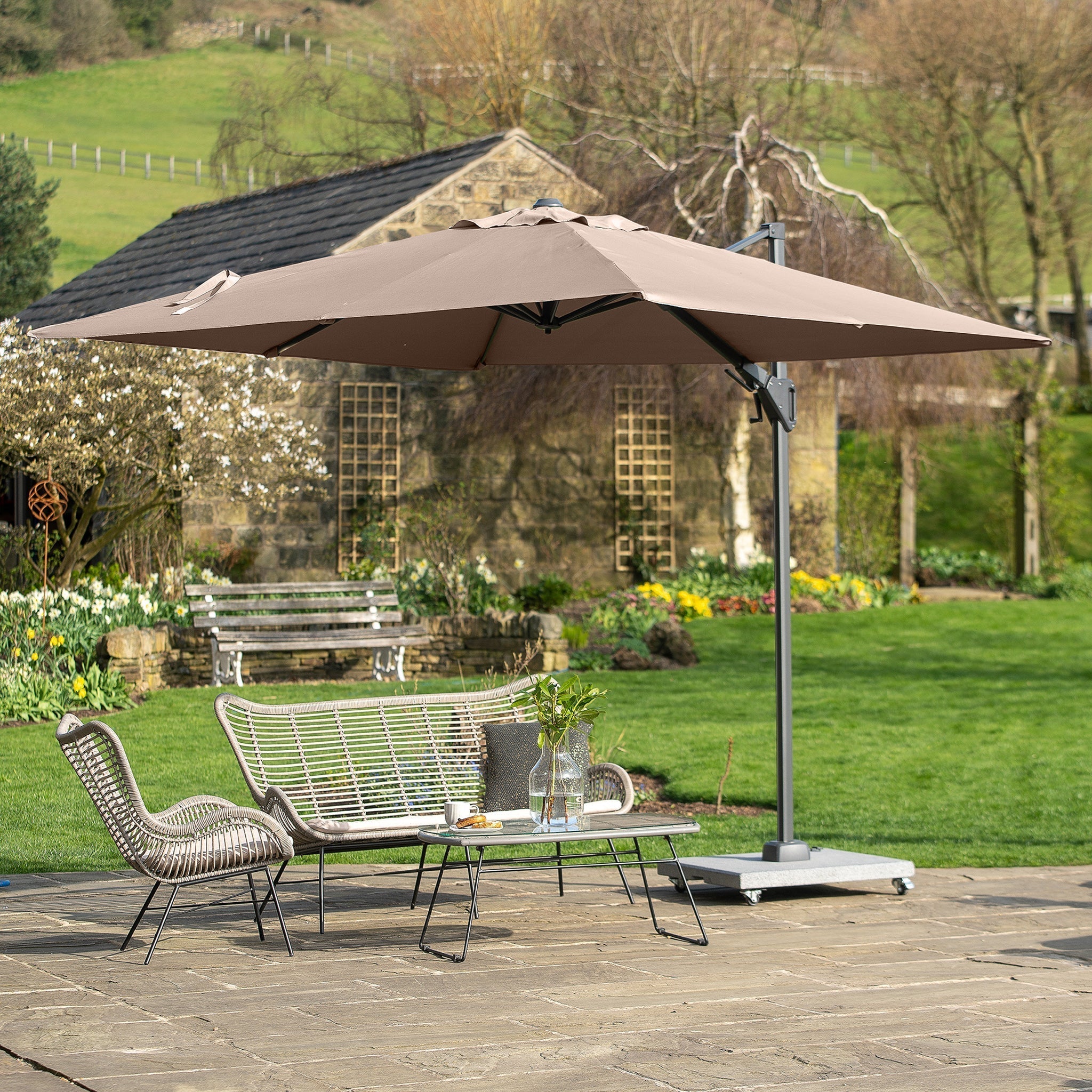 A patio with chairs, a bench, a table, and a Voyager T2 2.7m Square Parasol in Taupe boasting 360° tilt, set in a garden with a stone path and lush greenery.