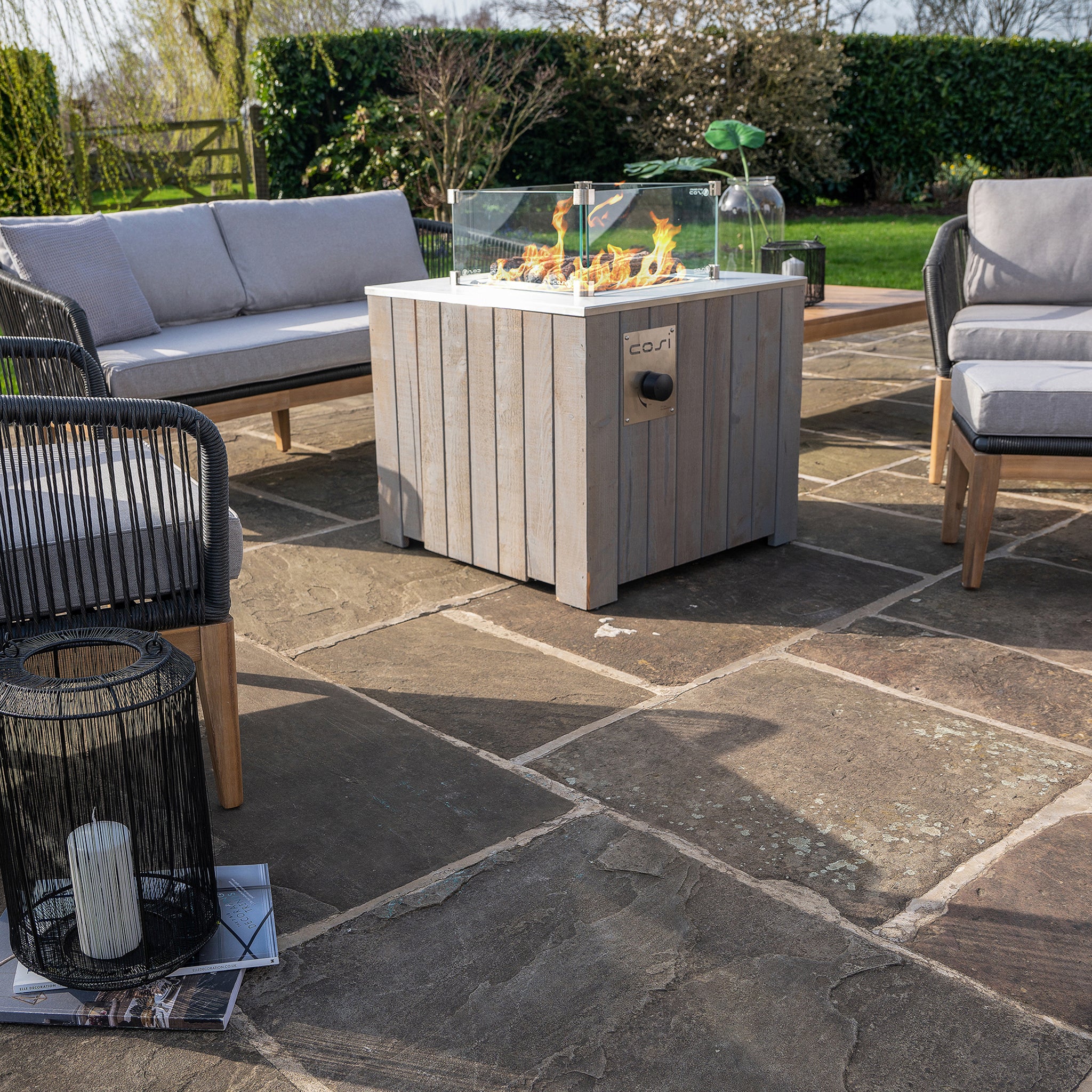 A patio with cushioned chairs and a Cosicube 70 Grey Wash Fire Pit surrounded by glass panels on stone flooring; green bushes in the background.