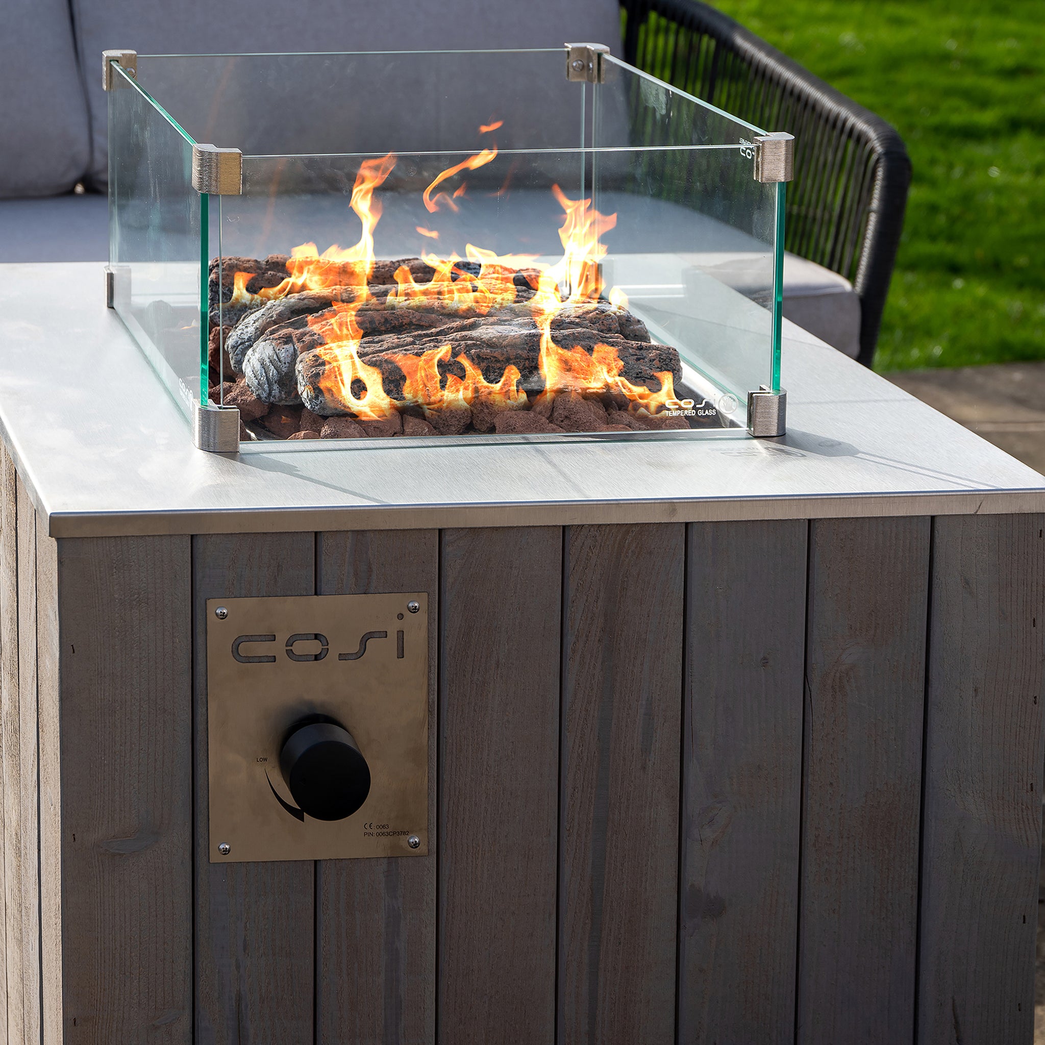 A modern outdoor fire pit, the Cosicube 70 Grey Wash Fire Pit, features a glass enclosure on a wooden table with flames burning inside, powered by an electronic ignition and fueled by a propane gas bottle.