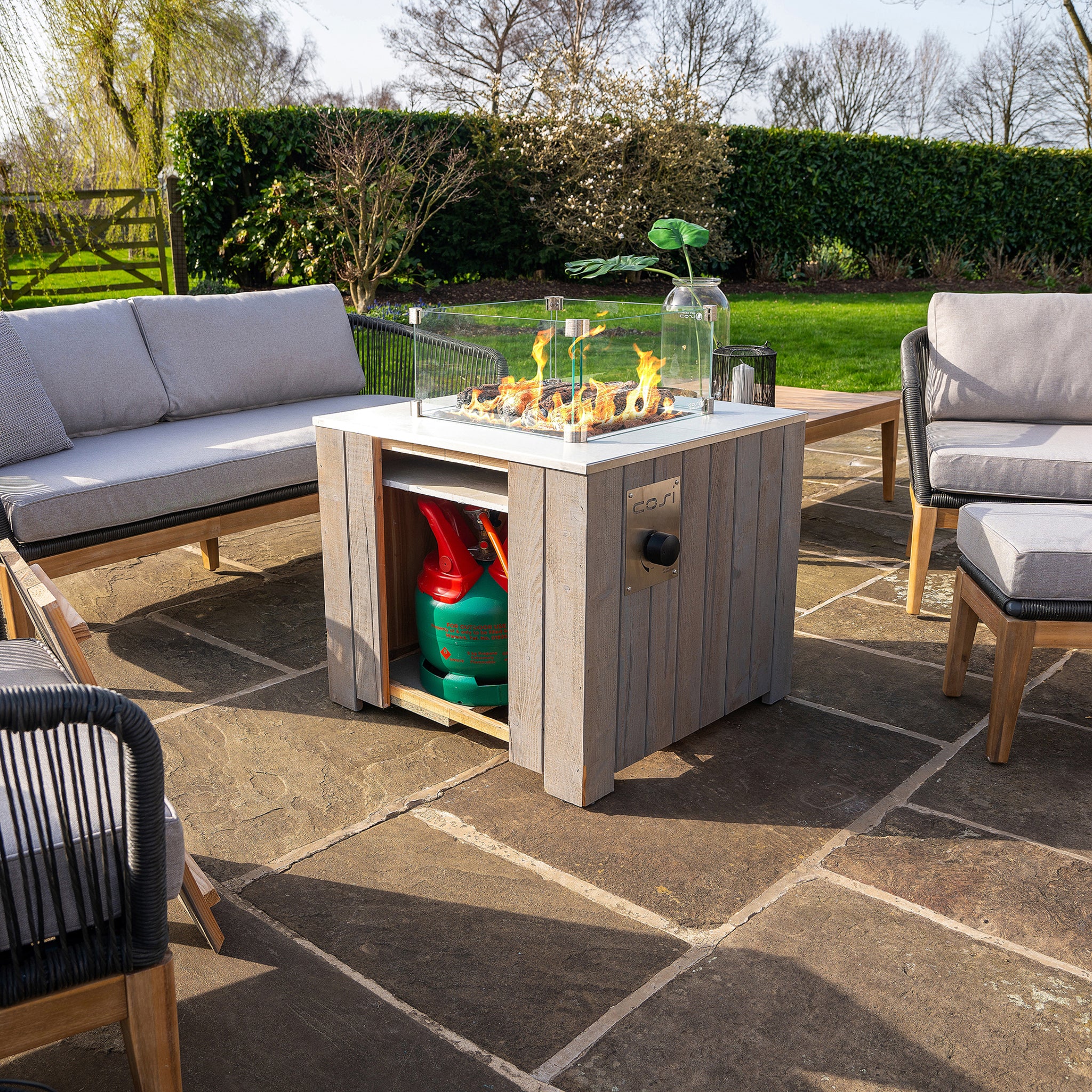 Outdoor patio featuring grey cushioned furniture, a Cosicube 70 Grey Wash Fire Pit with electronic ignition, and a view of a garden in the background.