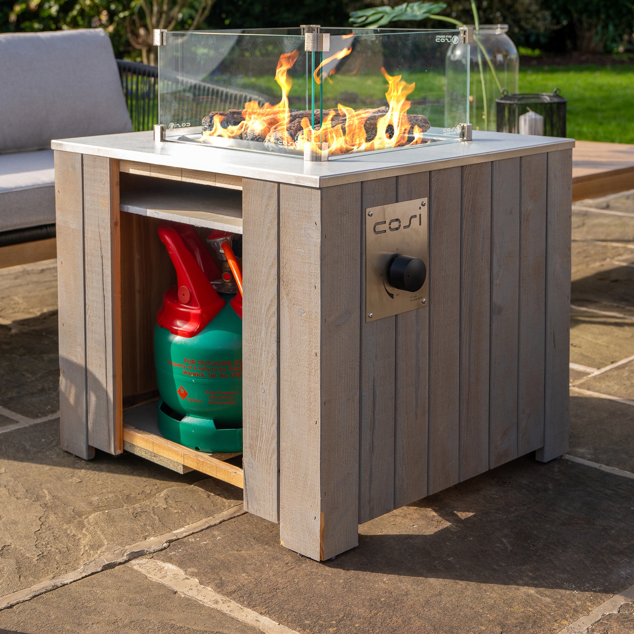 A Cosicube 70 Grey Wash Fire Pit with visible flames is placed on a stone patio. The storage below cleverly conceals a propane gas bottle and features an electronic ignition for easy lighting.