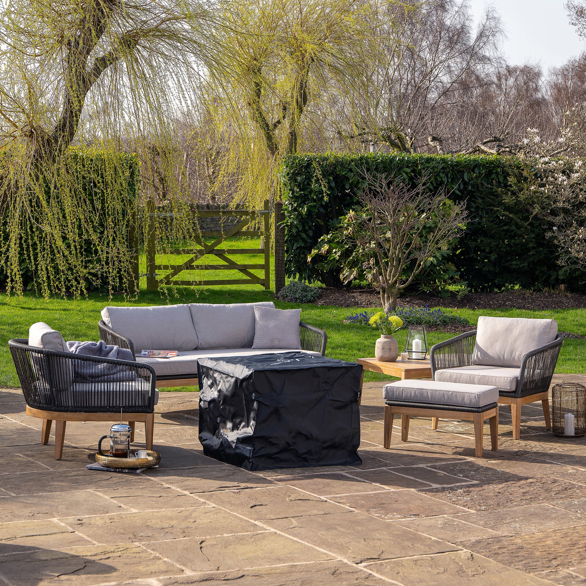 Outdoor patio with gray cushioned seating, a covered fire pit featuring electronic ignition, and a serene garden view with trees and a gated fence in the background. Enjoy cozy evenings around the stylish Cosicube 70 Grey Wash Fire Pit, easily fueled by a hidden propane gas bottle.