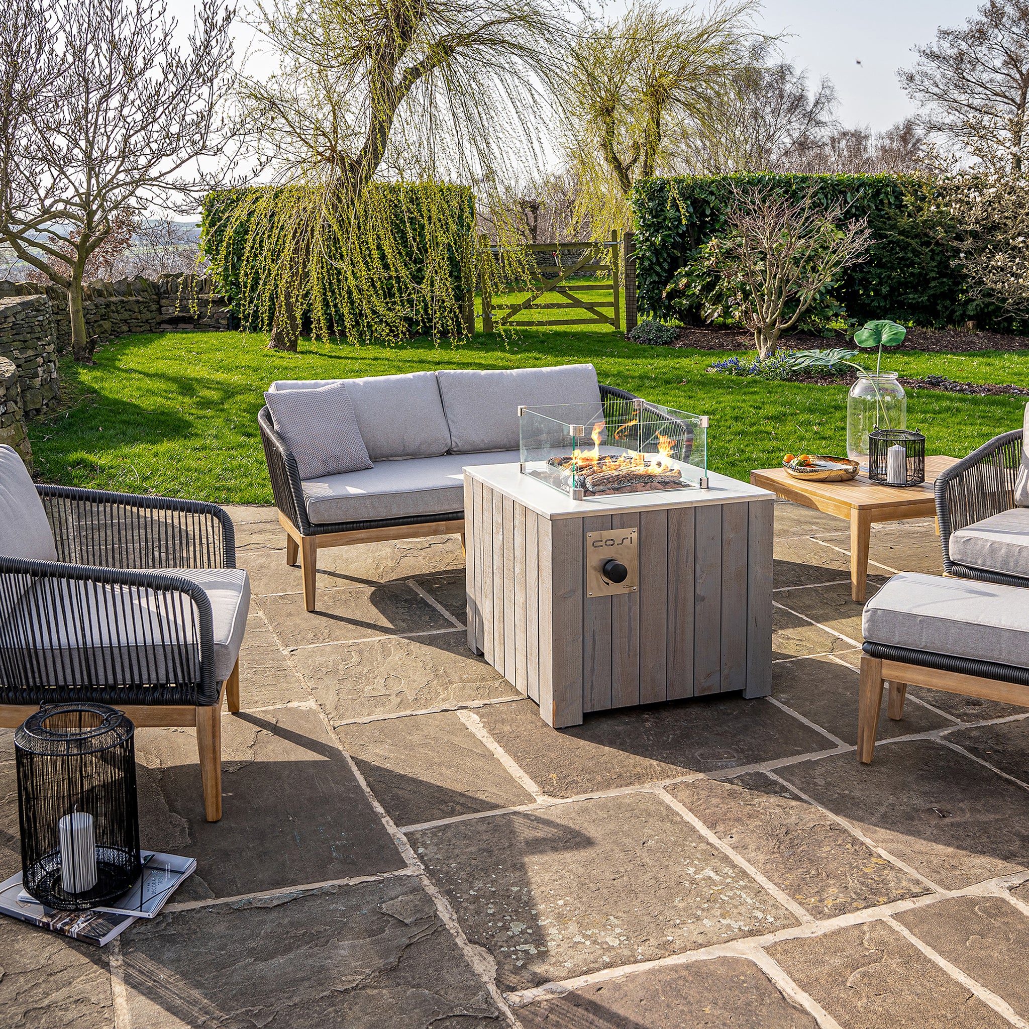 Outdoor patio featuring a Cosicube 70 Grey Wash Fire Pit, surrounded by a wicker furniture set. The serene retreat is enhanced by lush greenery and trees in the background, while the discreet propane gas bottle ensures effortless use with its electronic ignition system.