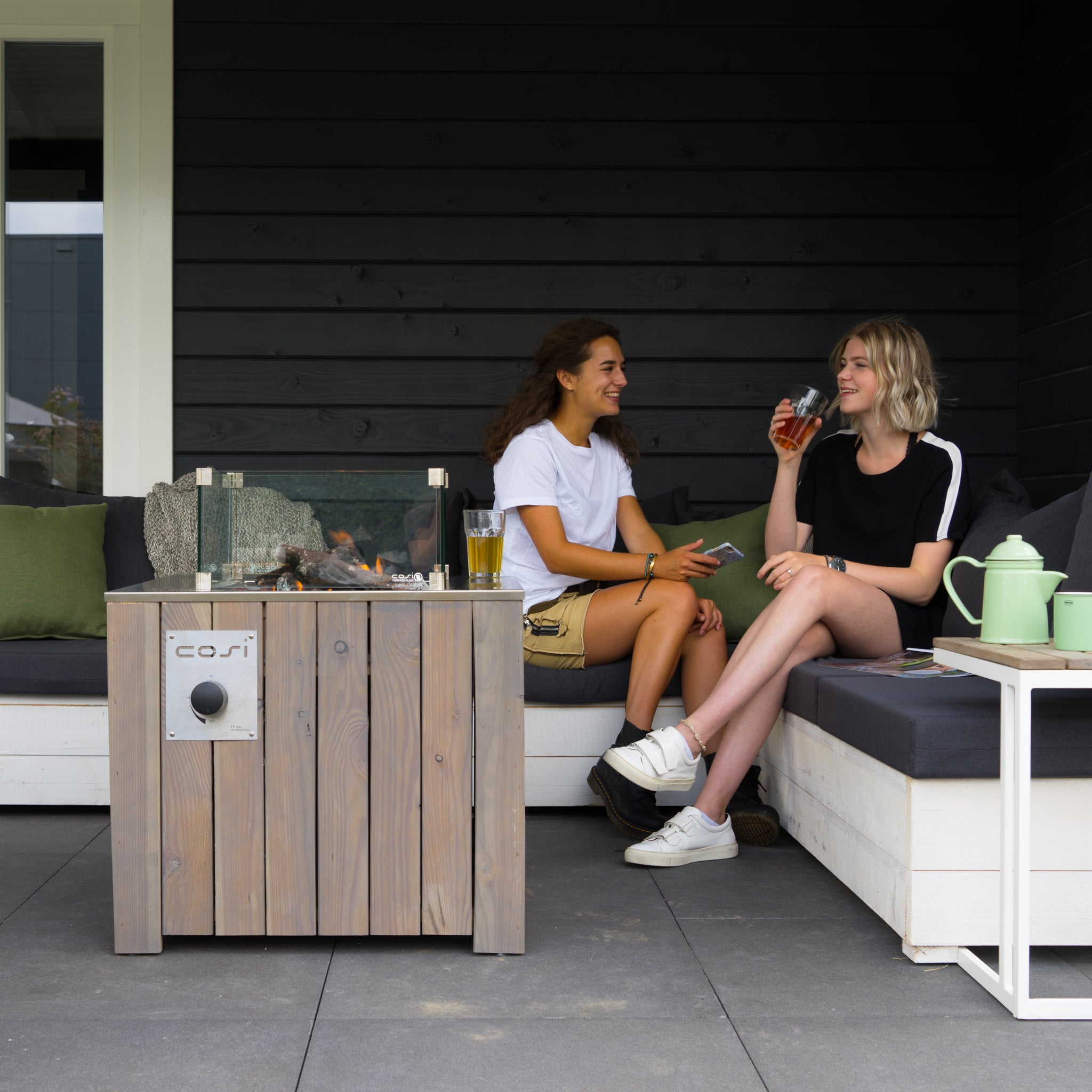 Two people sit and chat on an outdoor sofa beside a Cosicube 70 Grey Wash Fire Pit with electronic ignition on a patio. Drinks are on the table.