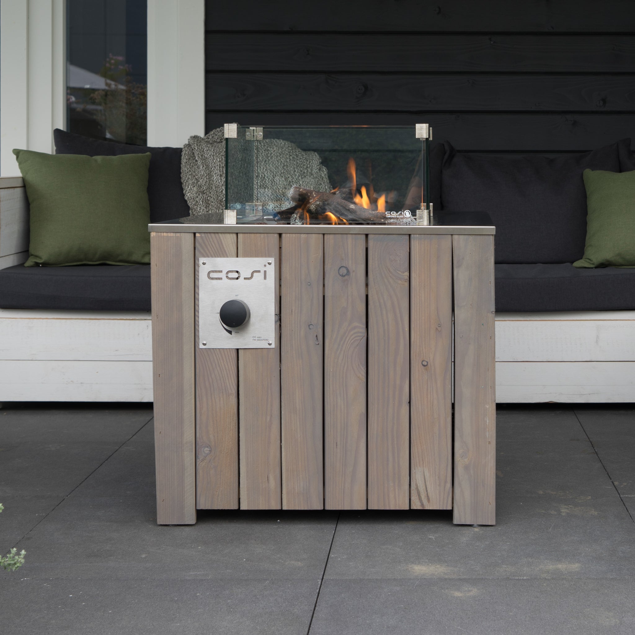 A Cosicube 70 Grey Wash Fire Pit with glass wind guards sits on the patio, complemented by a sofa with green cushions in the background. The fire pit features electronic ignition for convenience and can be connected to a propane gas bottle for constant flame enjoyment.