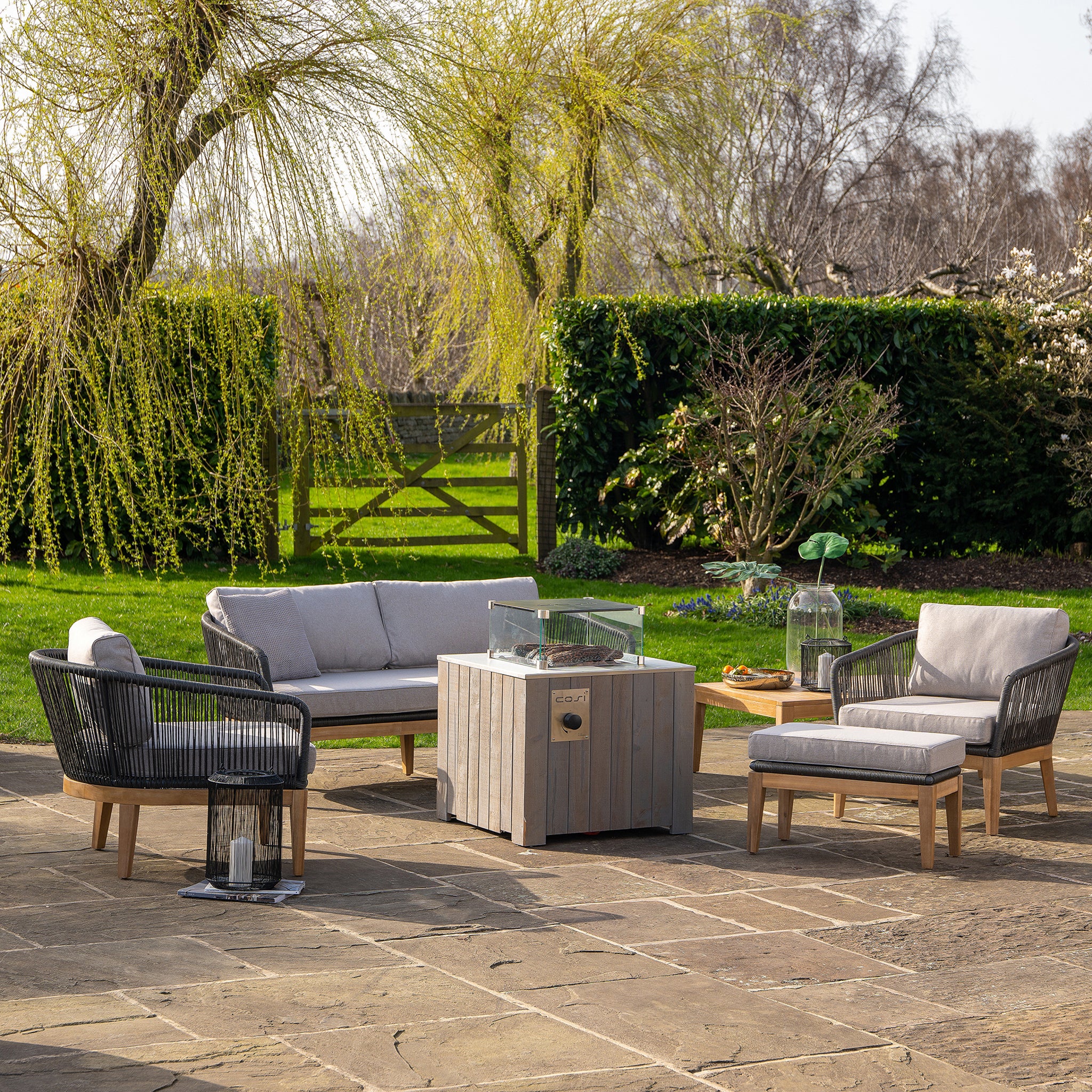 Outdoor patio with a sofa, chairs, and a Cosicube 70 Grey Wash Fire Pit featuring electronic ignition, set on a stone surface, surrounded by greenery and trees.