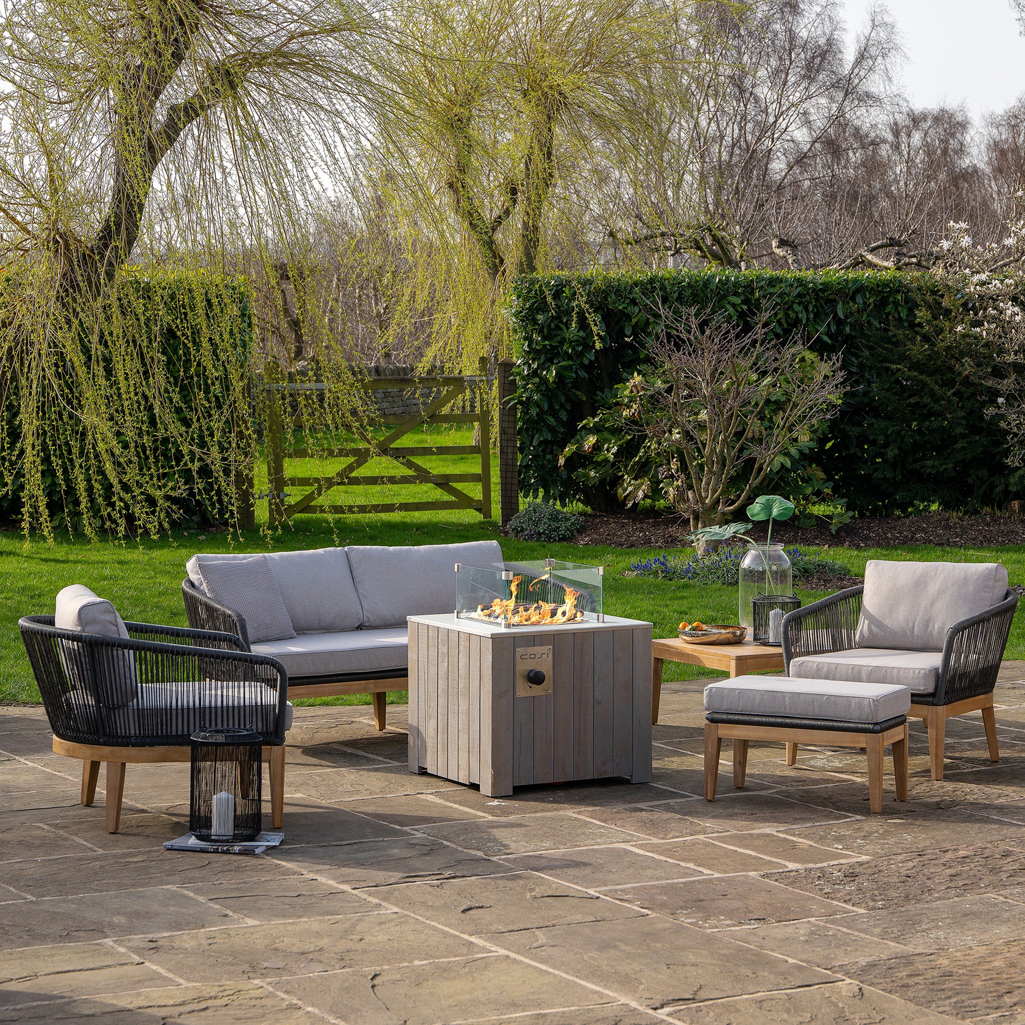 Outdoor patio with chairs, sofa, and Cosicube 70 Grey Wash Fire Pit featuring electronic ignition on stone flooring, set against a green lawn and trees in the background.