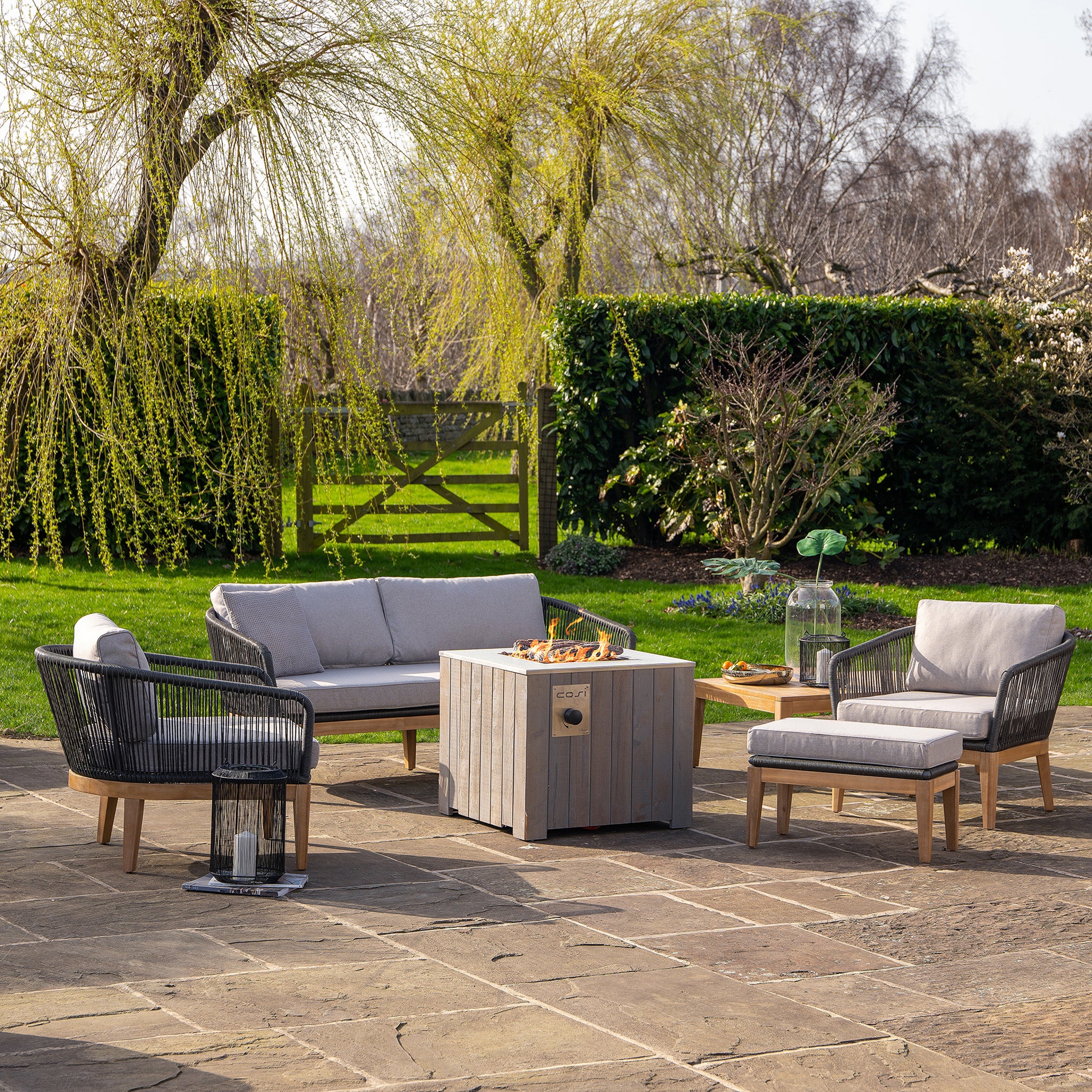 Outdoor patio with gray cushioned seating and a wooden fire pit table, featuring the Cosicube 70 Grey Wash Fire Pit with electronic ignition, surrounded by lush greenery and trees.